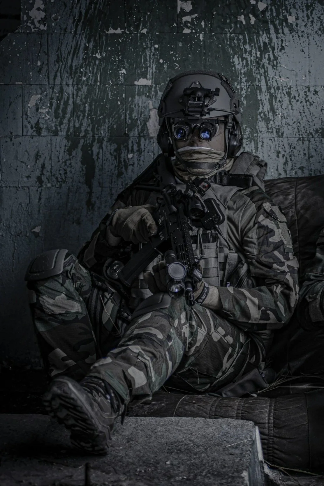 A soldier in camouflage uniform with tactical gear, helmet, and night vision goggles, holding a rifle, sitting against a dark, textured wall.