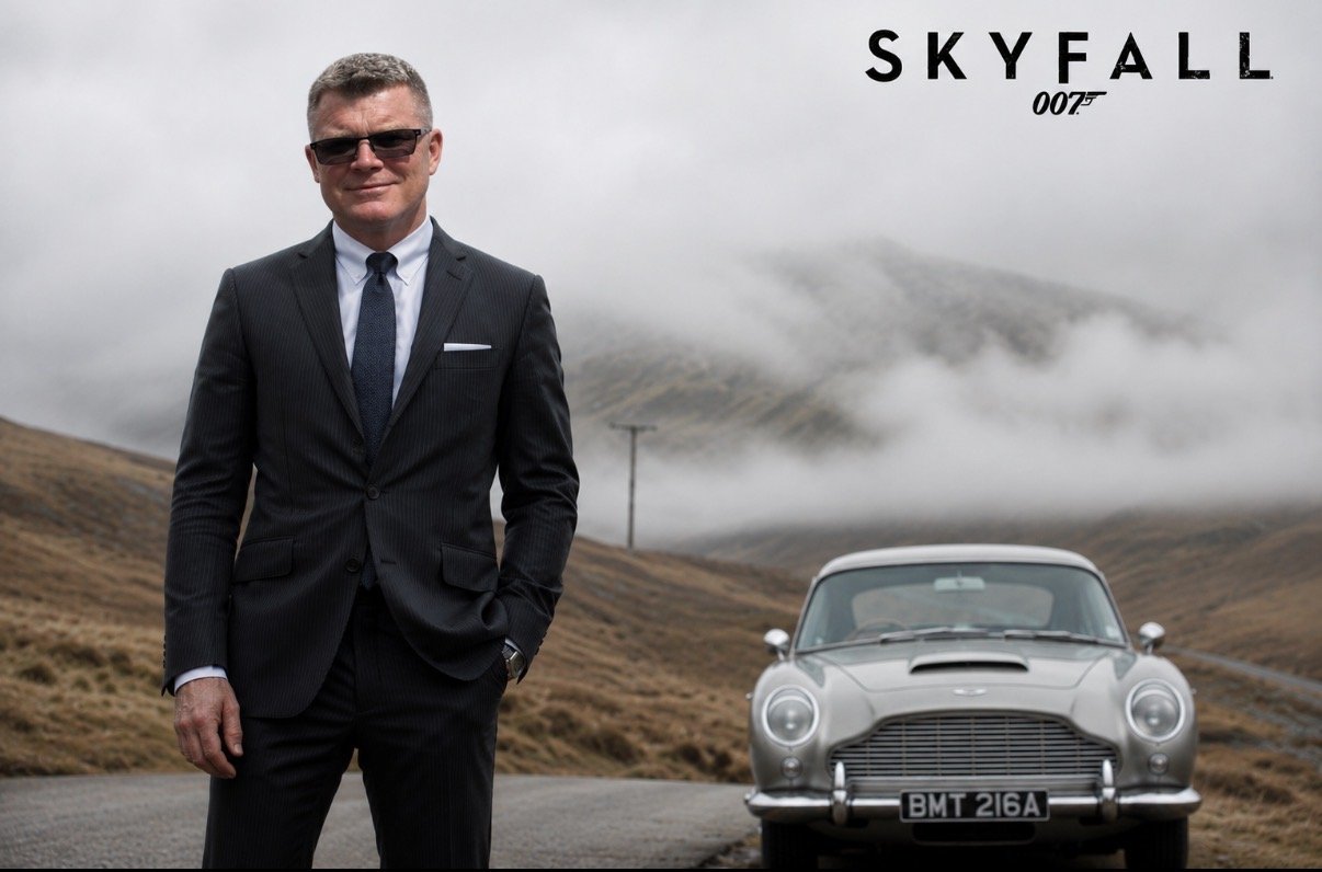 A man in a black pinstripe suit, sunglasses, and a tie stands outdoors with one hand in his pocket, smiling at the camera. Behind him, there is a classic silver Aston Martin car with a mountainous landscape and foggy sky. The 'Skyfall' logo and '007' emblem are in the top right corner.