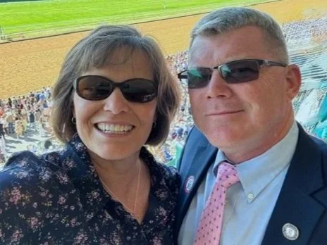 A smiling woman and man taking a selfie at a baseball game, with stadium seating and the field in the background.