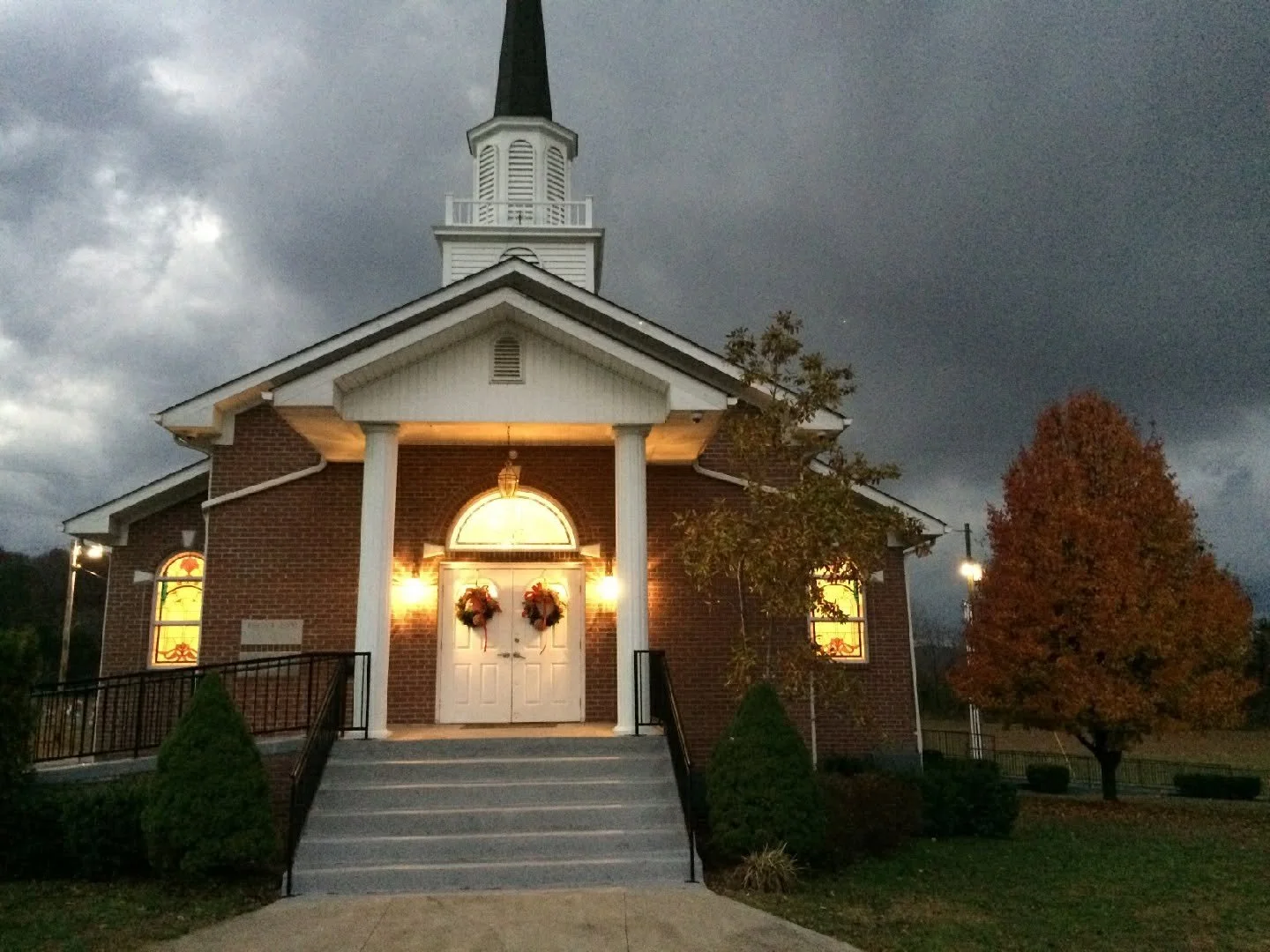 A small brick church with white accents, decorated for Christmas with wreaths on the door, illuminated by exterior lights, under dark stormy skies, and surrounded by trees and shrubbery.