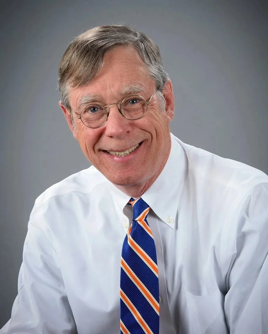 Portrait of Jeff Lowe with gray hair, glasses, smiling, wearing a white shirt and a blue, orange, and white striped tie, against a gray background.