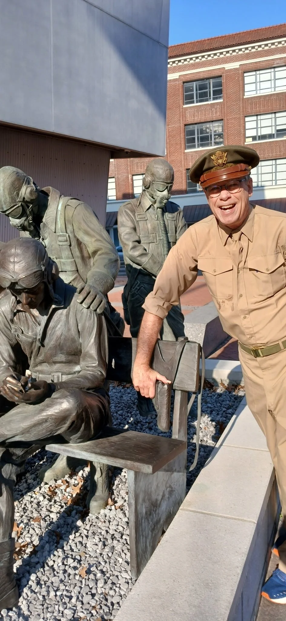 A man in a beige uniform and military hat smiling and posing next to bronze statues of soldiers, one seated and two standing, with a modern building and blue sky in the background.