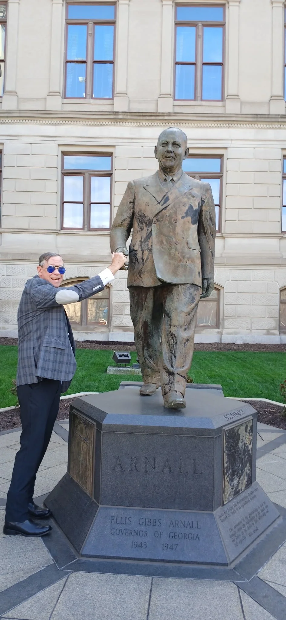 A man in sunglasses and a plaid blazer is smiling and touching the hand of a statue of Ellis Gibbs Arnall, the former governor of Georgia, in front of a historic building. The statue is made of bronze and depicts a man in a suit walking with a confident stride. The base of the statue has inscriptions, including Ellis Gibbs Arnall's name and his tenure as governor from 1943 to 1947.