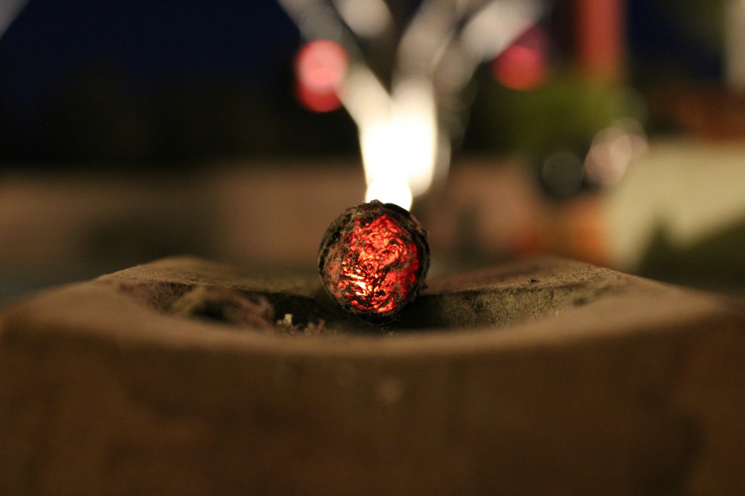 A close-up of an actively burning cigar glowing red at the tip, resting on an ashtray in a low light setting. Sold at Outlaw Vapor in Preston and Malad, Idaho.