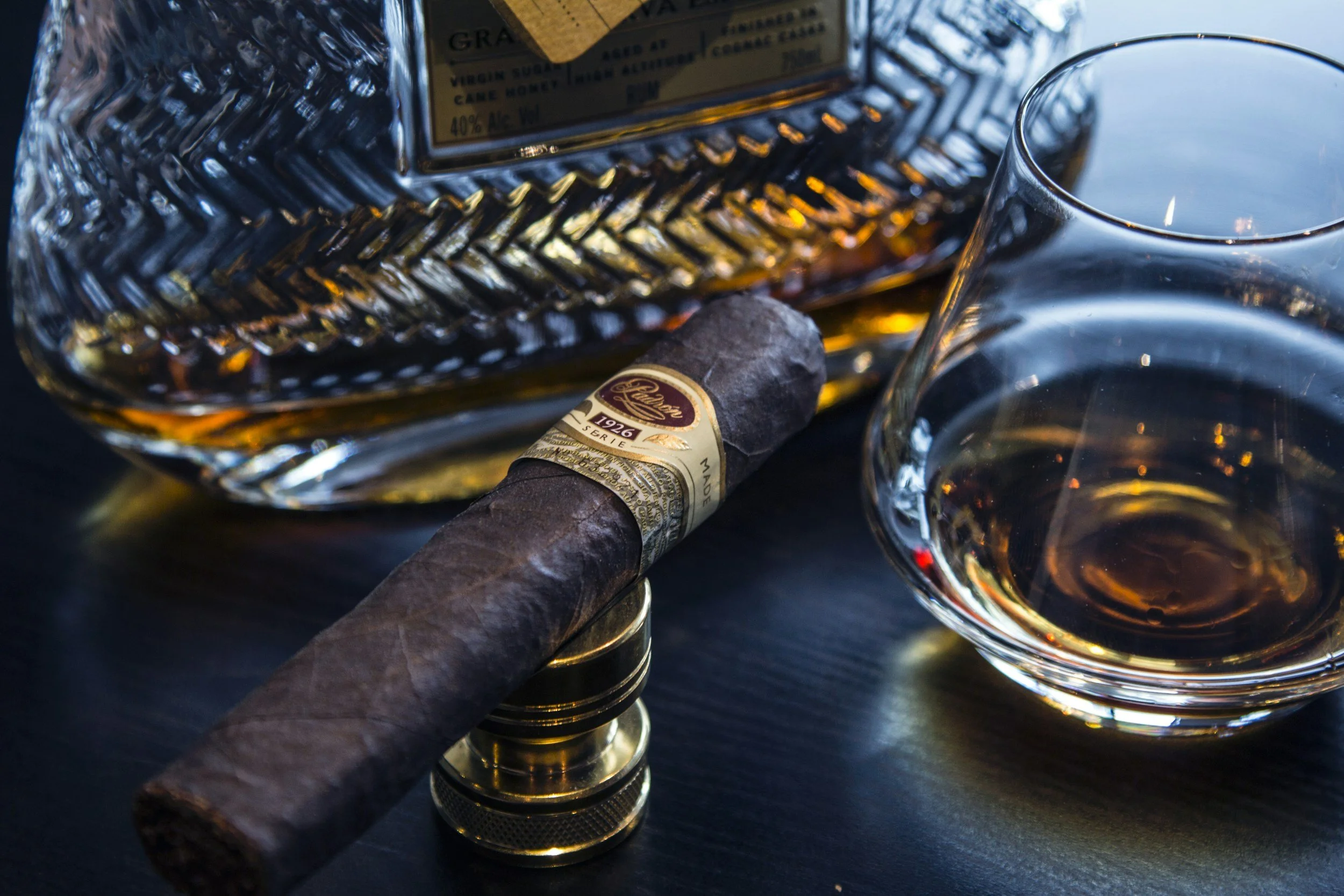 A glass of whiskey, a PERDOMO cigar set on a cigar holder, a bottle of aged bourbon on a dark surface.  Sold at Outlaw Vapor in Preston and Malad, Idaho.