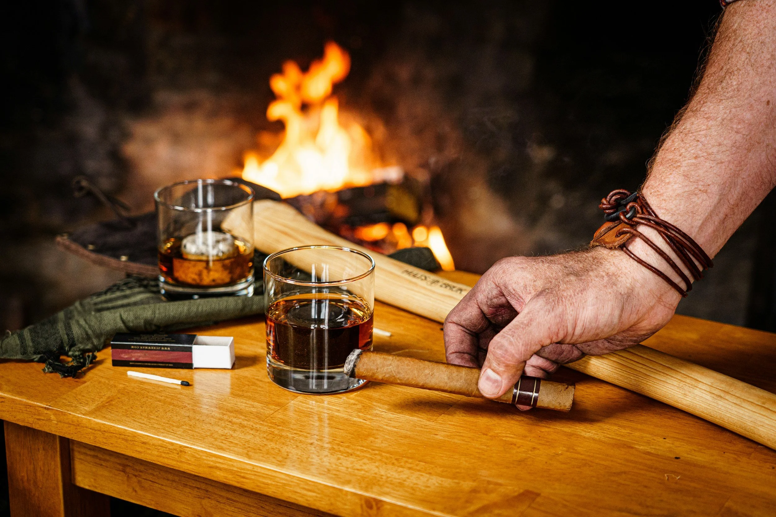 A mans right arm is holding a PERDOMO cigar in a masculine setting with a fire lit, a glass of bourbon, and an axe.  Sold at Outlaw Vapor in Preston and Malad, Idaho.