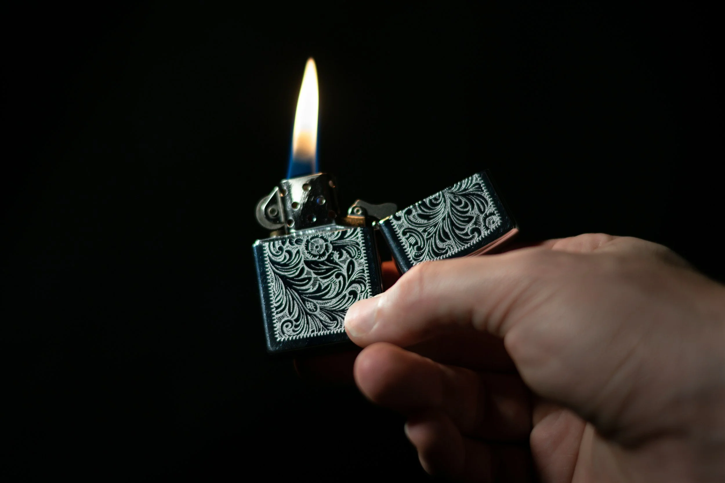 A person's hand holding a decorated silver Zippo lighter with a flame against a black background. Sold at Outlaw Vapor in Preston and Malad, Idaho.