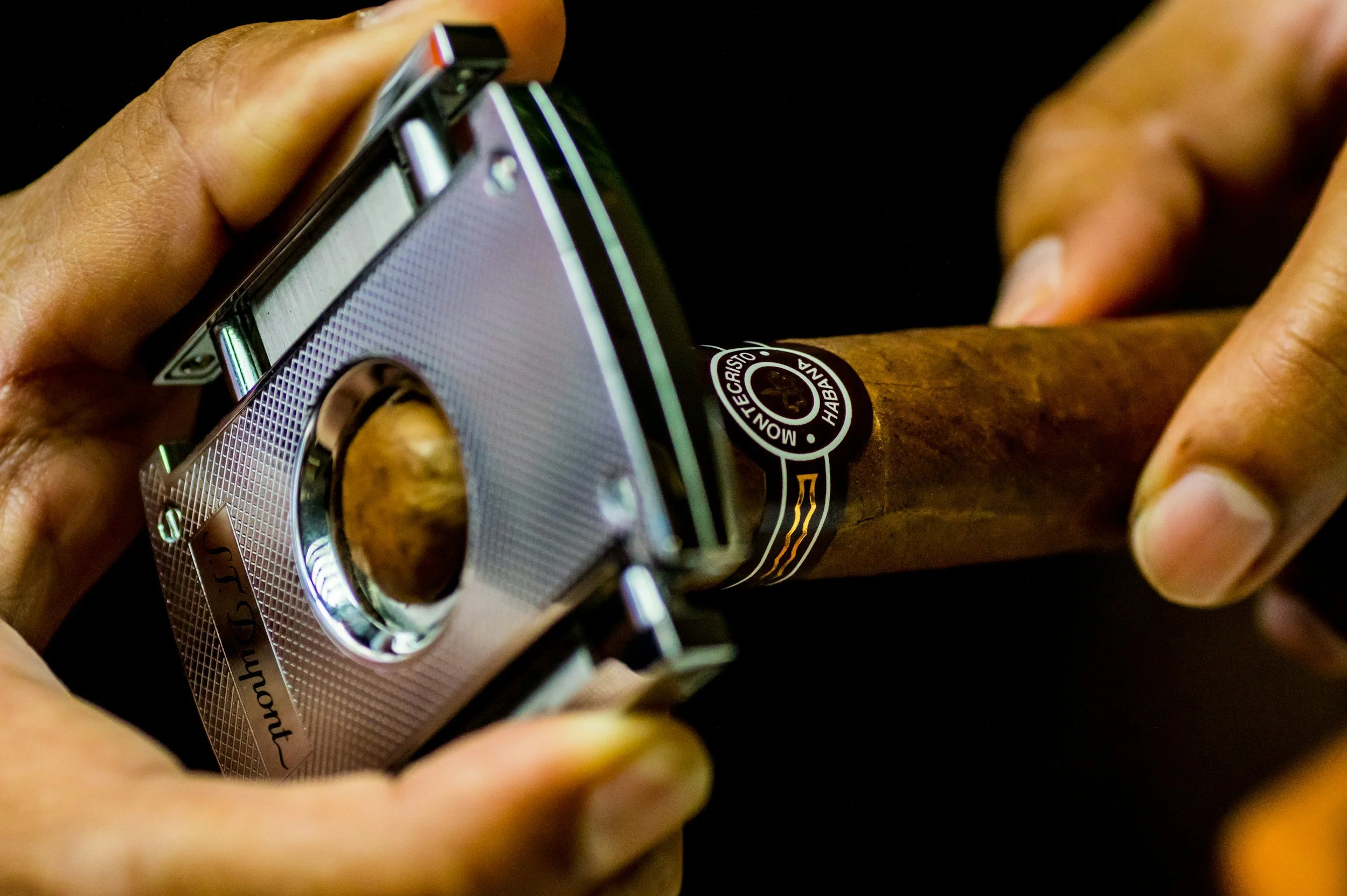 Close-up of hands holding a cigar cutter as it prepares to clip the tip of a cigar with a band that reads 'HABANA'. Sold at Outlaw Vapor in Preston and Malad, Idaho.