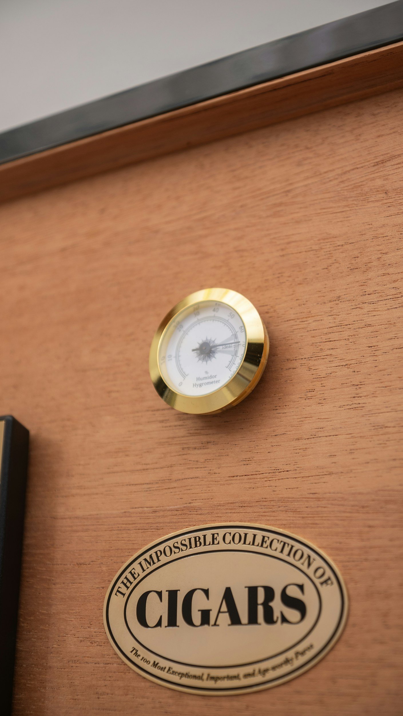 Close-up of a wooden cigar humidor box with a round humidity gauge and a label reading 'The Impossible Collection of Cigars'. Sold at Outlaw Vapor in Preston and Malad, Idaho.