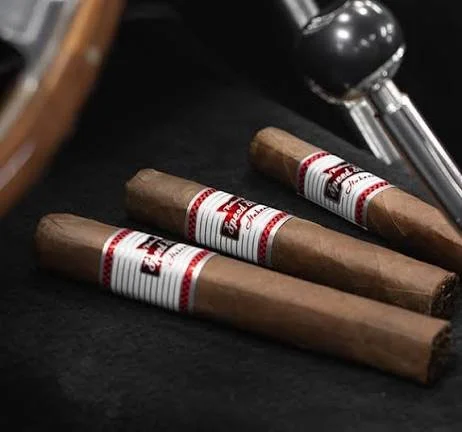 Three CAO cigars with white and red bands lying on a black surface near a metal lighter. Sold at Outlaw Vapor in Preston and Malad, Idaho.