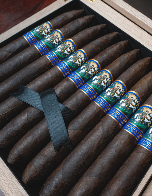 A wooden box containing ten FOUNDATION cigars with blue and gold bands labeled 'Fundador.' The cigars are dark brown and are held together with a black ribbon. Sold at Outlaw Vapor in Preston and Malad, Idaho.