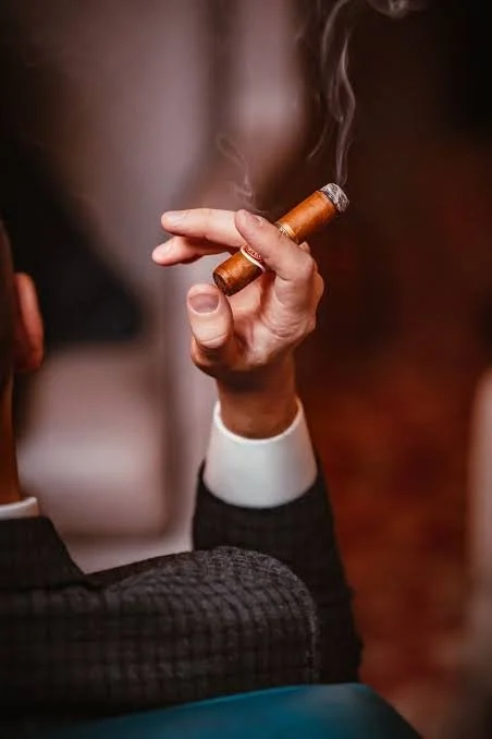 A gentleman sitting in a cigar lounge, holding a Drew Estates Cigar in his right hand. The cigar is lit with light smoke coming from the end of the cigar.  Sold at Outlaw Vapor in Preston and Malad, Idaho.