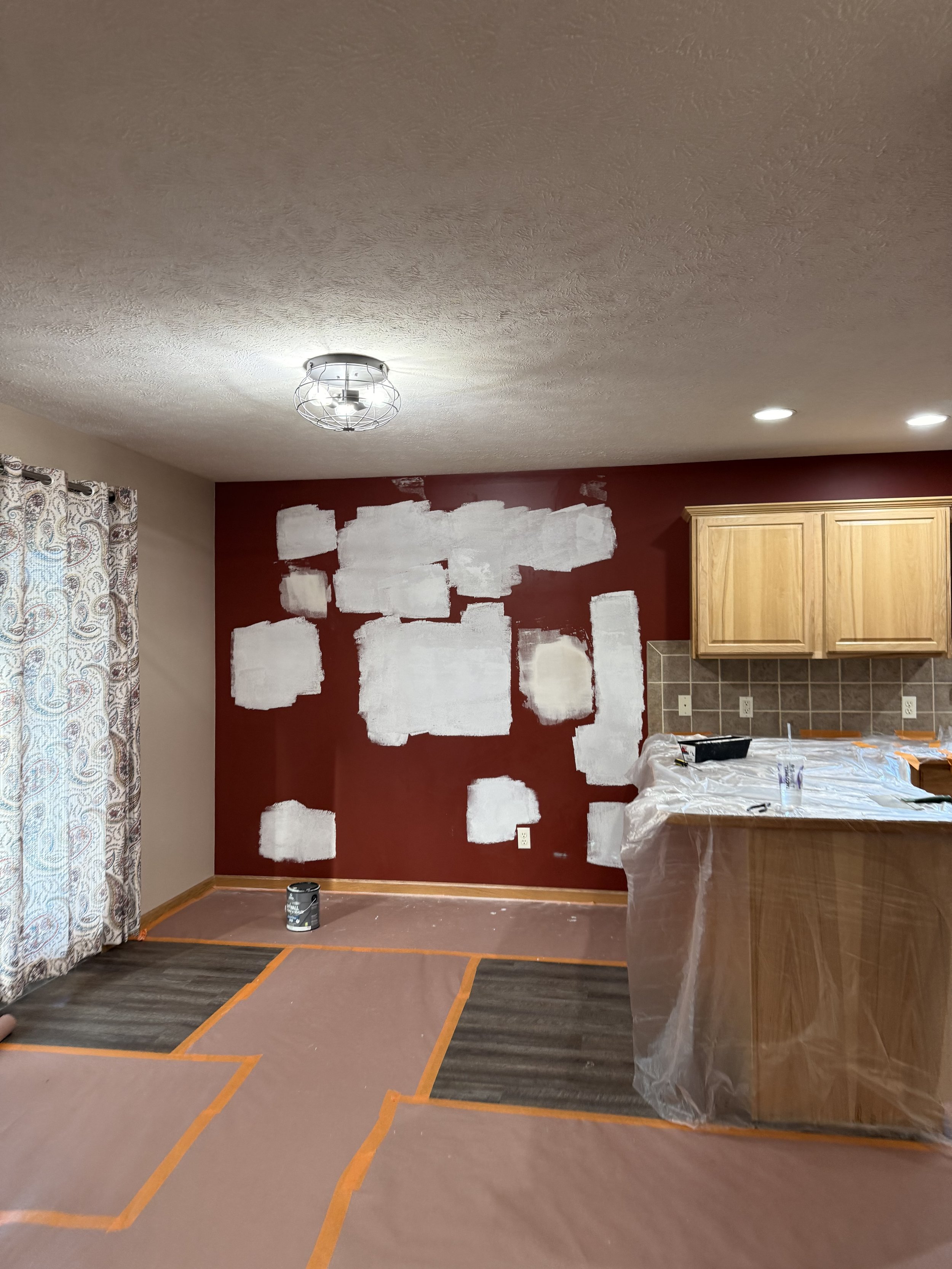 Living room area during painting renovation, with a red accent wall partially painted white, a ceiling light, and a kitchen cabinets.