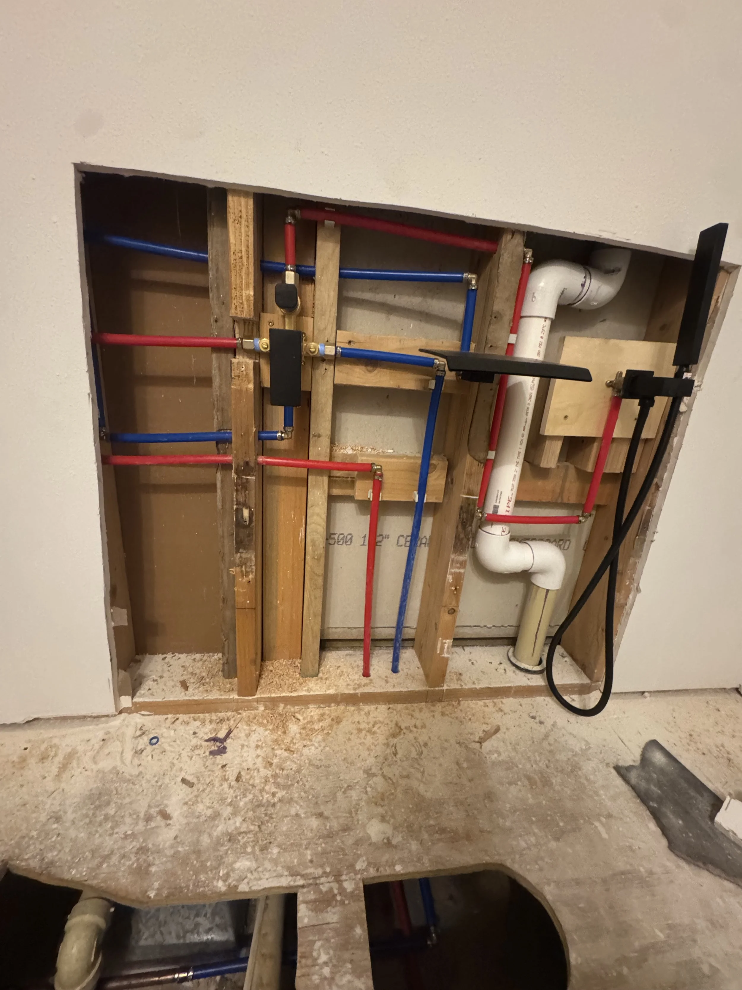 Partial view of a plumbing installation with red and blue water pipes, PVC pipe, and wooden framing in a ceiling opening under construction.