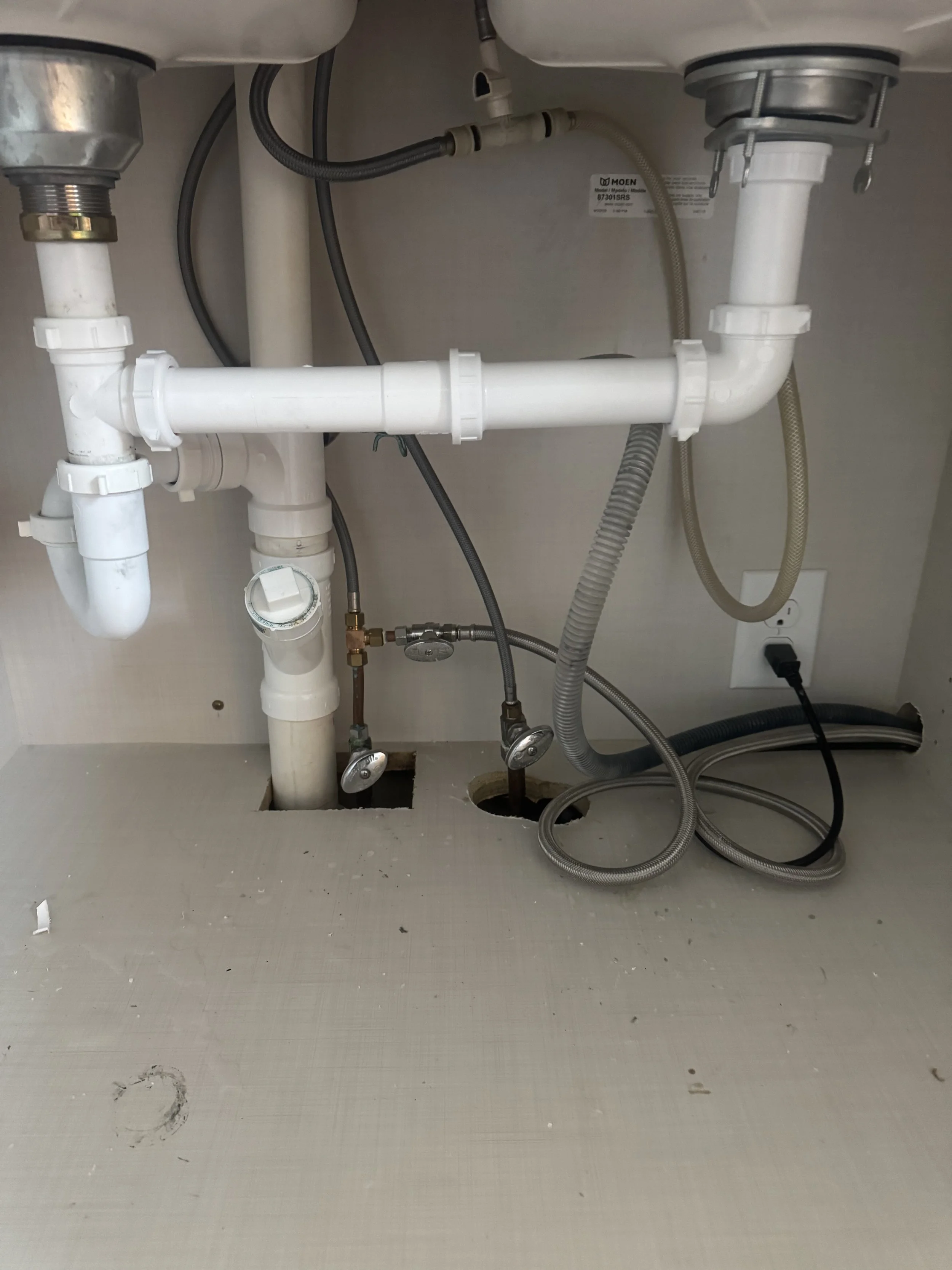 Underneath a kitchen sink, showing white PVC pipes, metal pipes, hoses, and a wall outlet, with some holes in the cabinet floor.