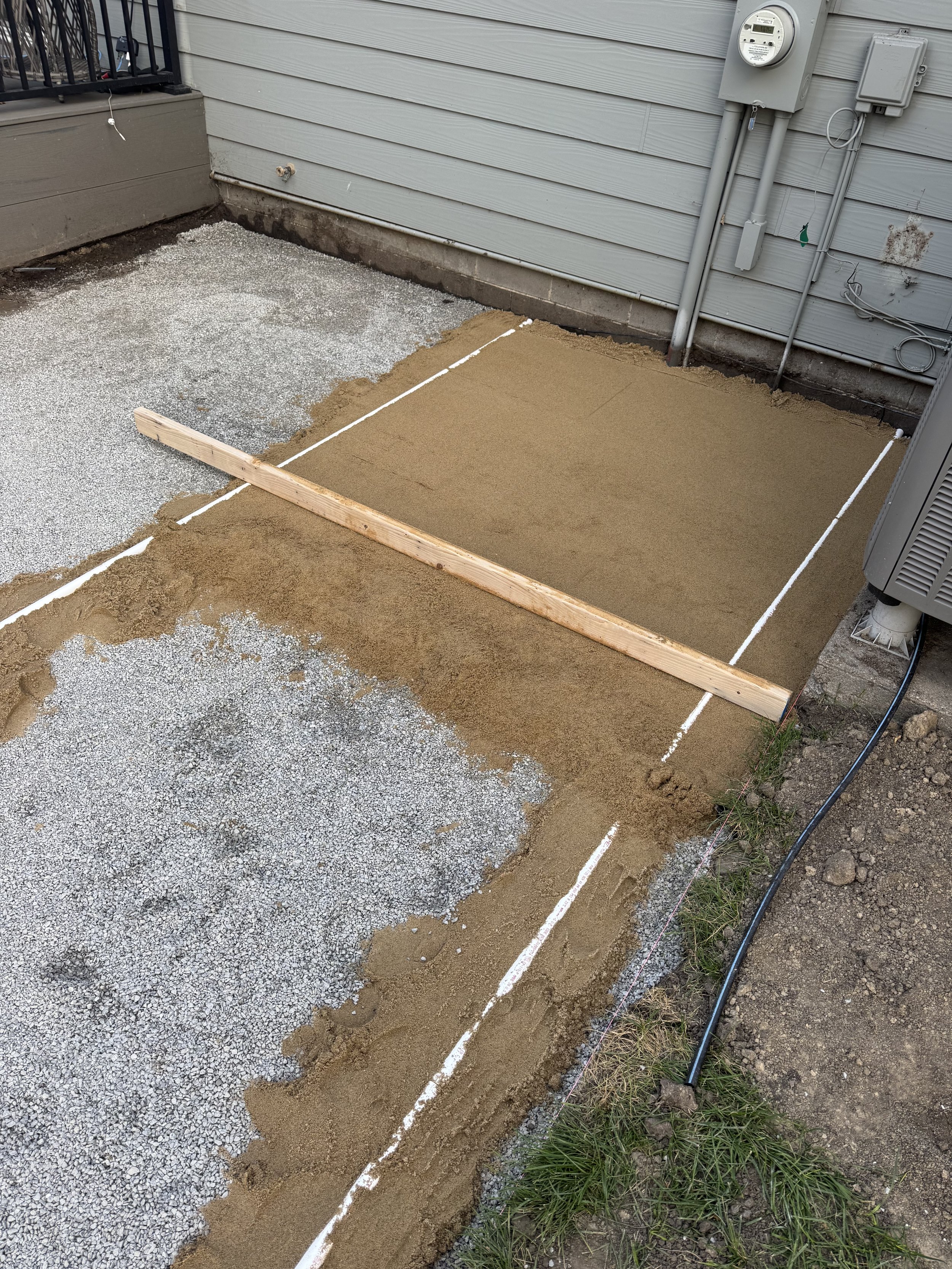 A construction site with a partially poured concrete slab and decorative sand or gravel, marked with white lines and a wooden board, alongside a house with visible siding, electrical meters, and outdoor wiring.