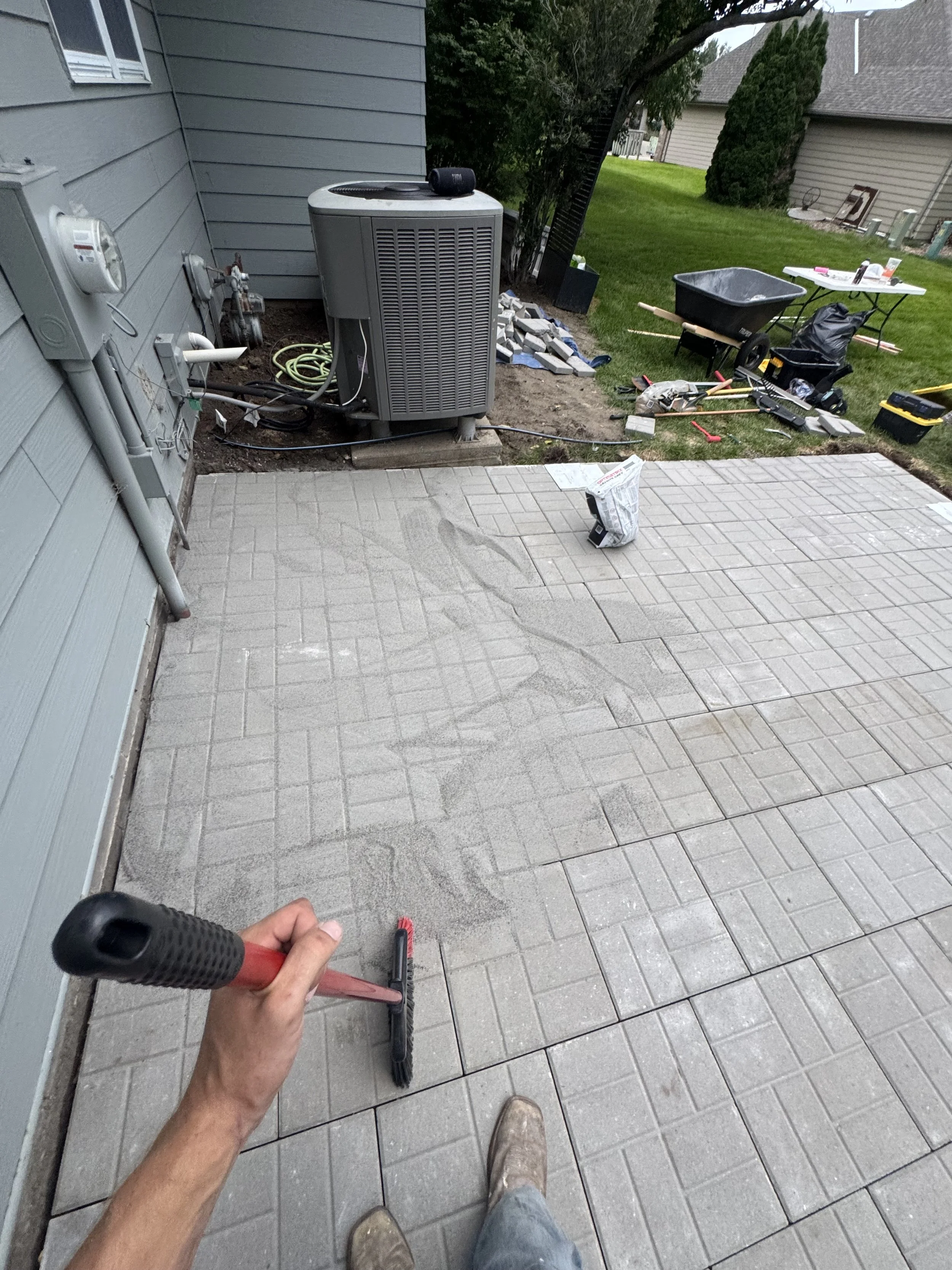 Person holding a red push broom on a light gray tiled porch, with a grassy yard, a wheelbarrow, various tools, and construction materials nearby.