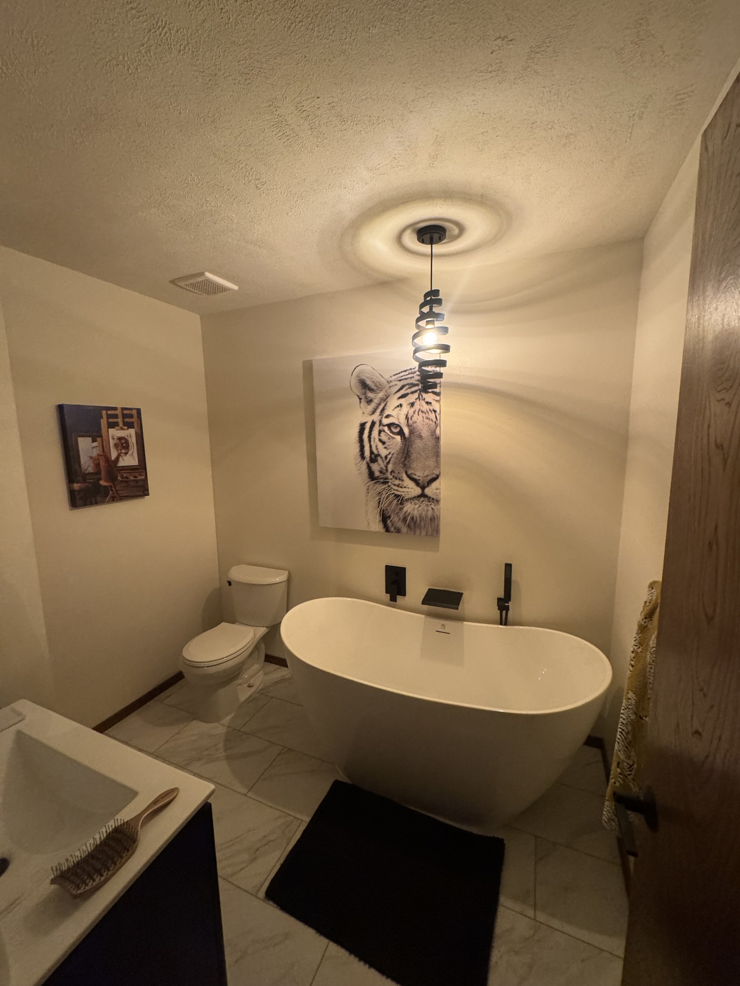 A bathroom with a modern freestanding bathtub, a portrait of a tiger on the wall, a toilet, and a small black rug on the tiled floor.