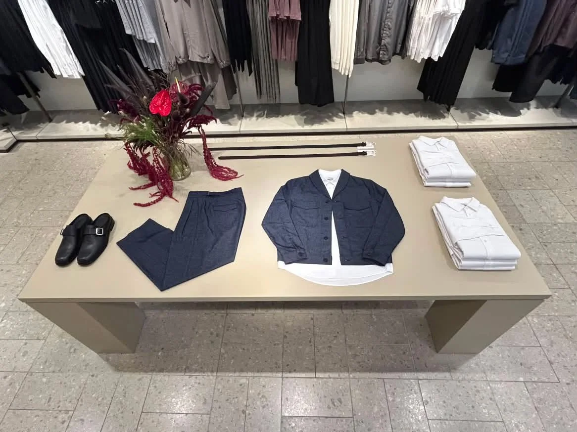 Display table with folded white shirts, navy pants, a navy jacket over a white shirt, black shoes, a large floral arrangement, and hanging clothes in the background.