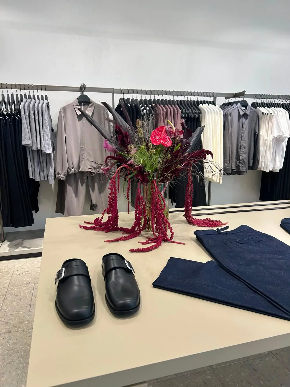 Clothing display with a floral arrangement on a table in a retail store. The table has folded jeans and black shoes. In the background, there are racks of various colored shirts and jackets.