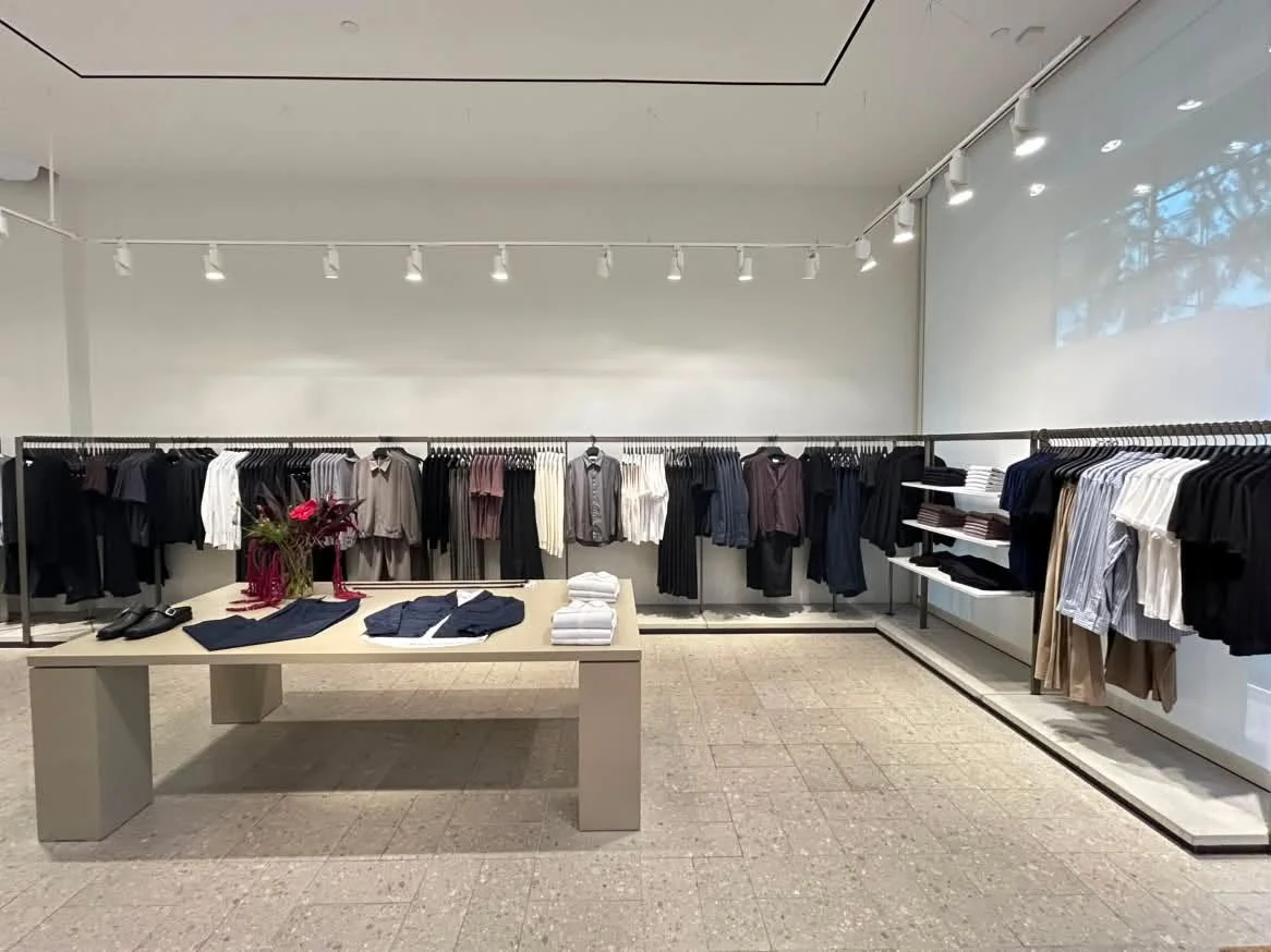 Clothing store display with shirts and jackets on racks and folded on shelves, table with neatly folded clothes, and a vase with flowers.