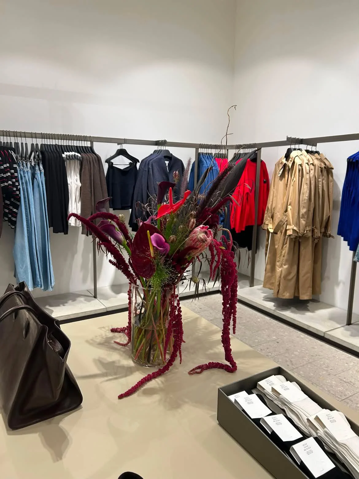 A retail clothing store display featuring a large floral arrangement in a glass vase on a table, with handbags nearby, and clothing racks with various jackets and pants in the background.