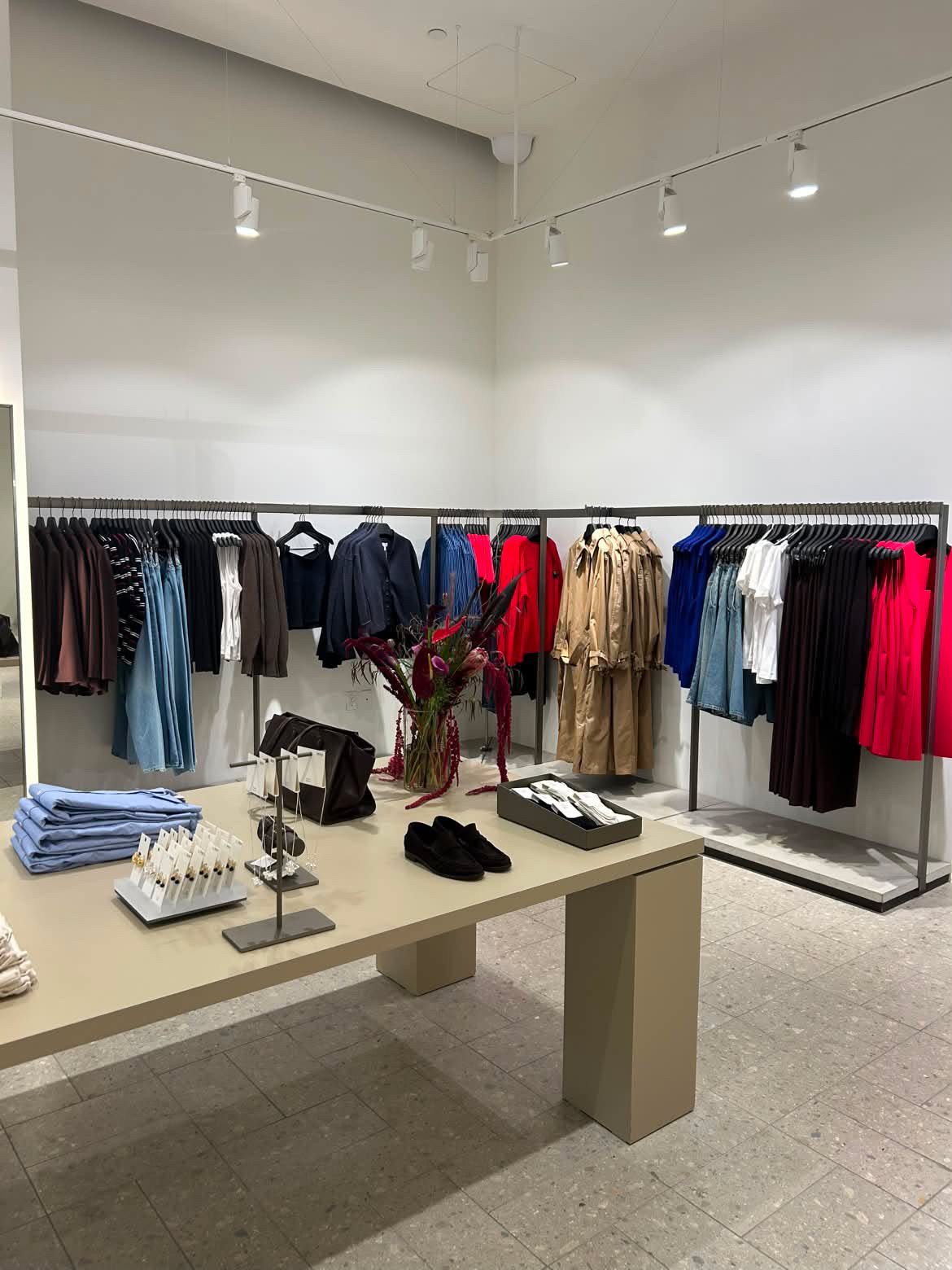 Clothing store display with racks of garments, a table with folded clothing, jewelry, shoes, and a floral arrangement in the center.