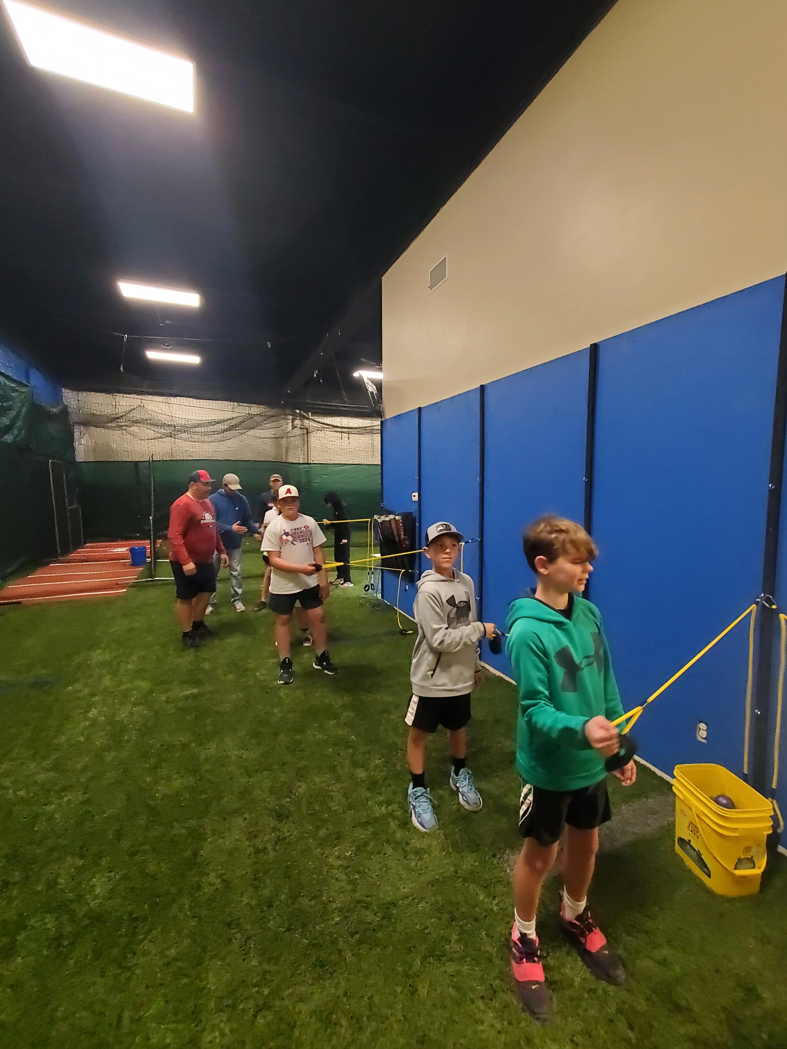 Indoor sports facility with children and teenagers standing in line holding golf balls and sticks next to a yellow bucket, with netted fencing and blue and beige walls.