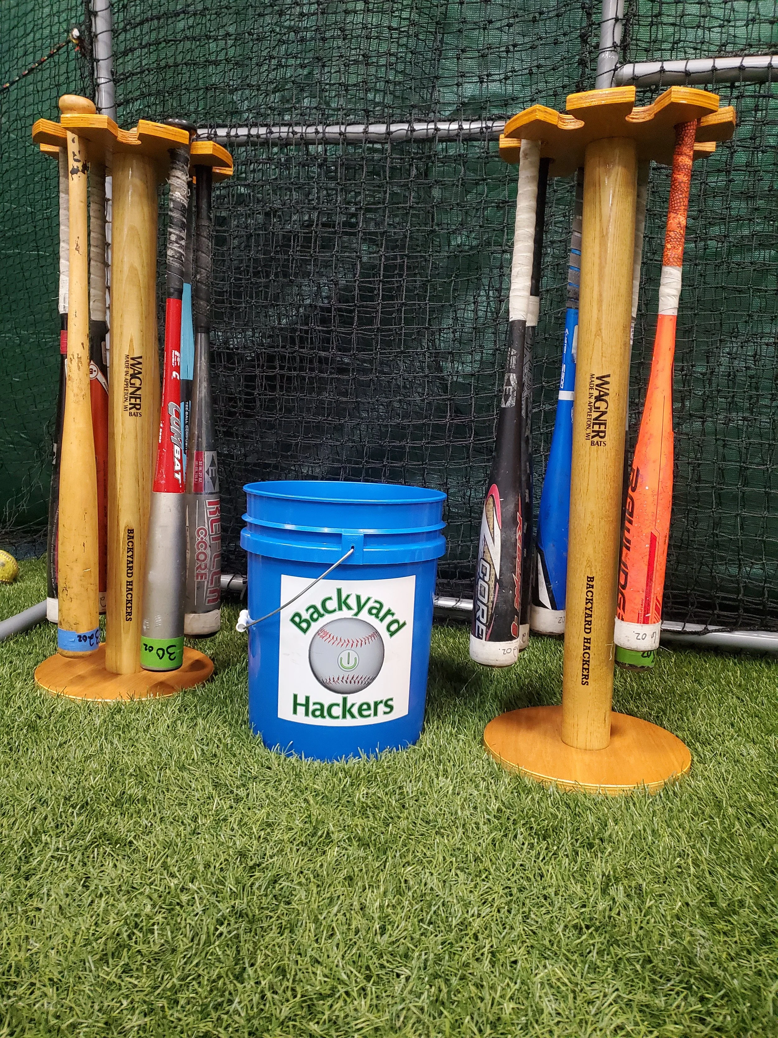 Row of six baseball bats resting on two wooden stands with a blue bucket labeled "Backyard Hackers" in front, all on artificial grass in front of a black netting backstop.