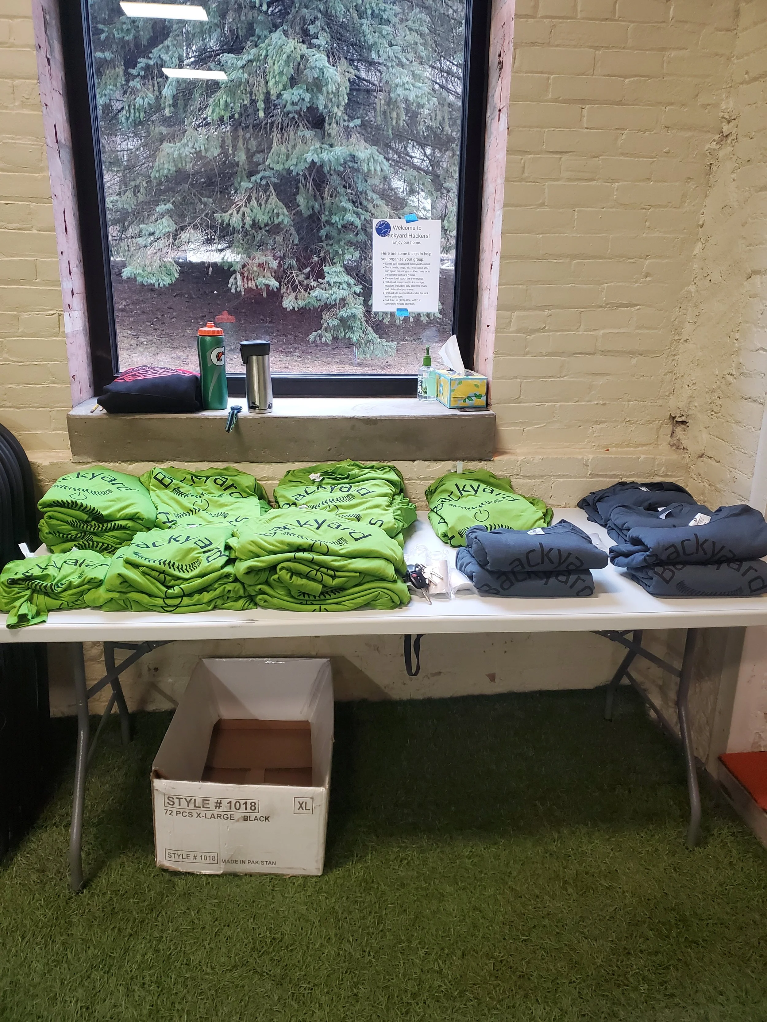 A table holding several green and gray folded t-shirts with the word 'Backyard' printed on them, a box of 72 XL black t-shirts, a box of tissue, a green spray bottle, and two thermoses. Behind the table, there is a window with a view of a large tree outside and a note taped to the window.