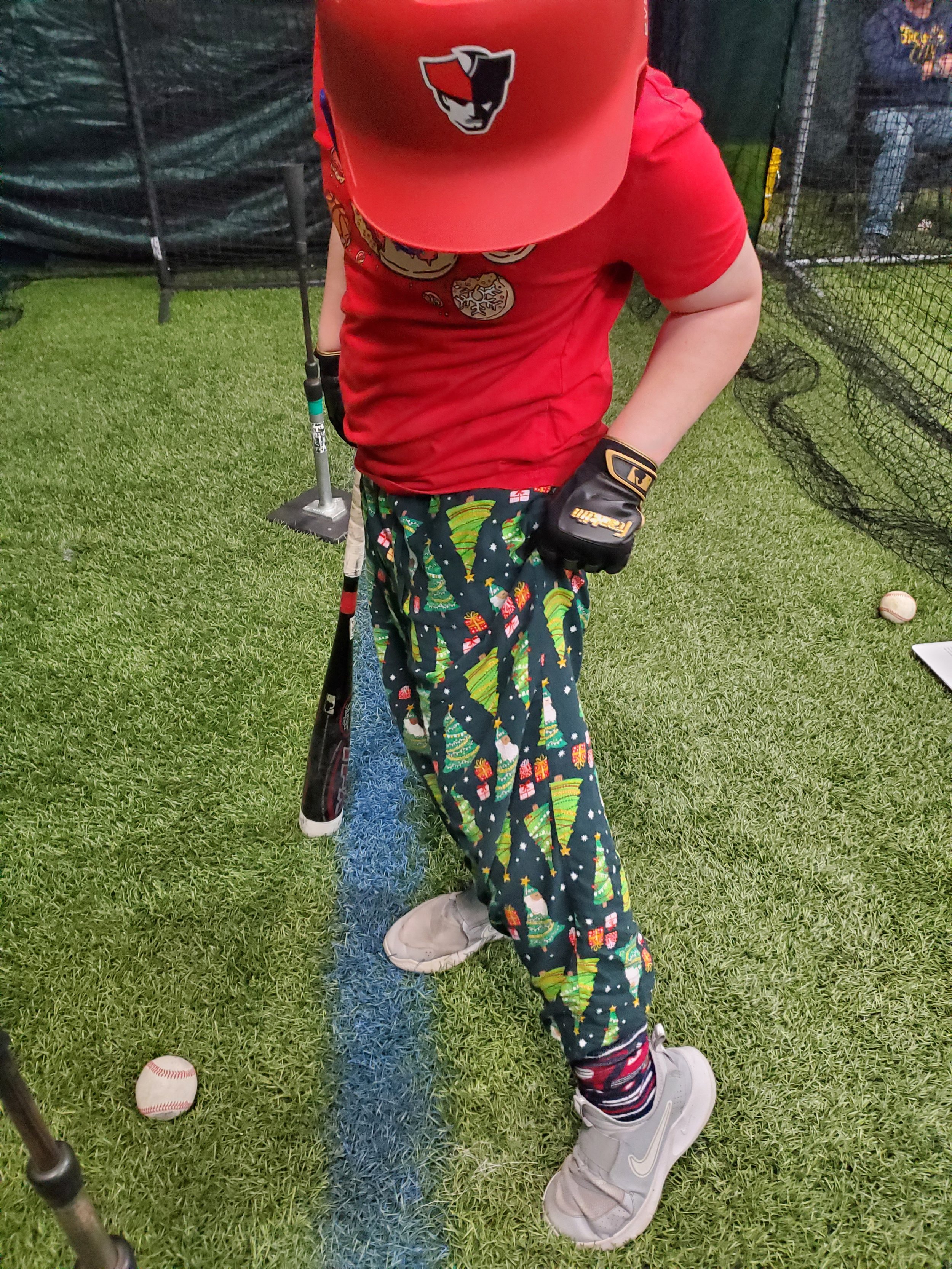 Close-up of a young boy in a red shirt and colorful Christmas pajama pants, wearing a red baseball cap with a black and white logo. The boy is holding a baseball bat in a batting stance on a grassy field, with two baseballs nearby.