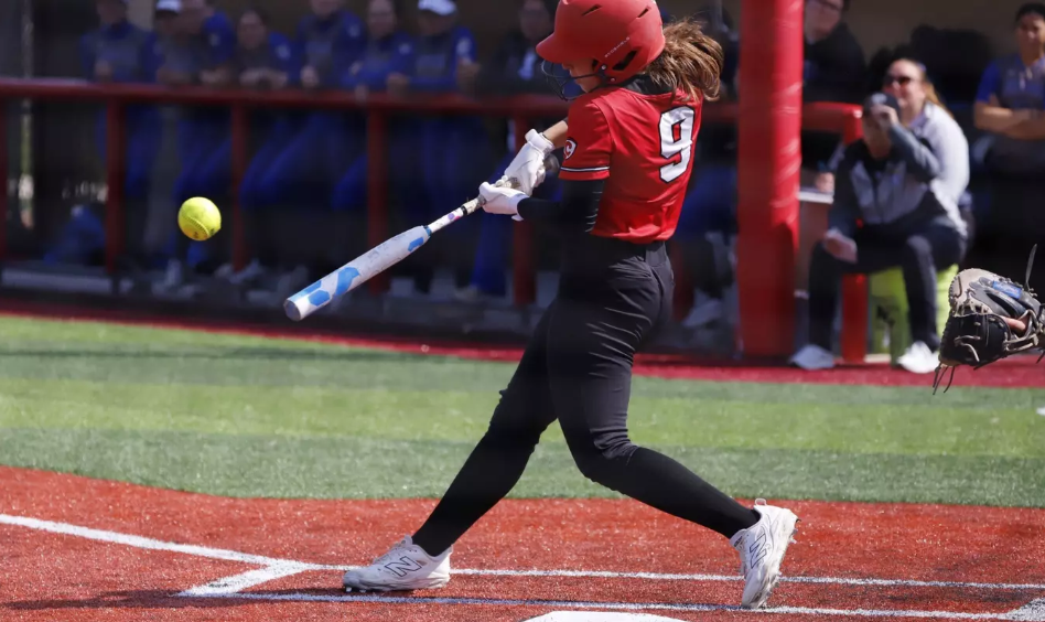 A young girl in a red softball uniform and helmet is swinging a bat at a yellow softball during a game.