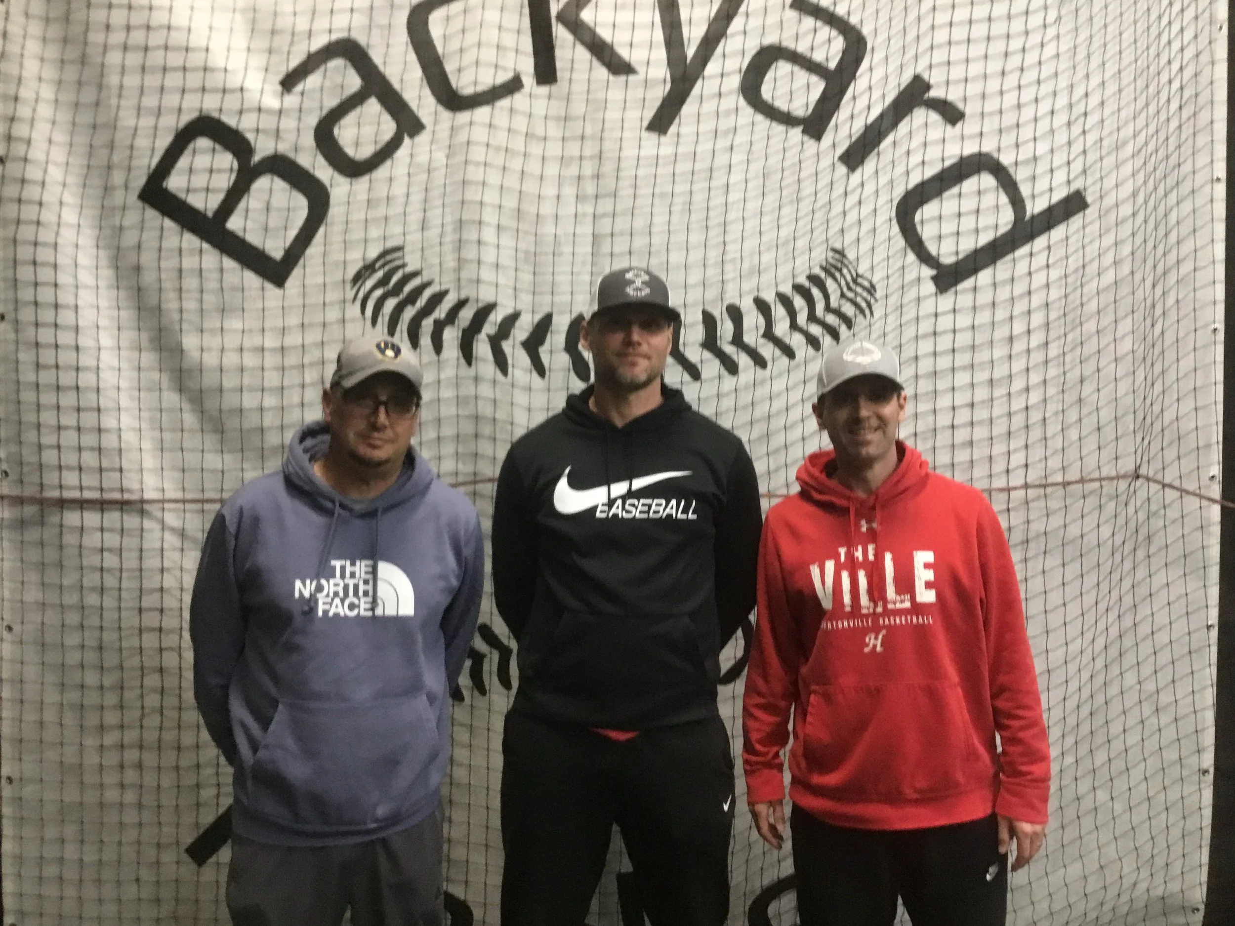Three men standing in front of a sports net with a large baseball-themed banner behind them. The man on the left is wearing a gray hoodie and glasses, the man in the center is wearing a black Nike hoodie, and the man on the right is wearing a red hoodie with white text.