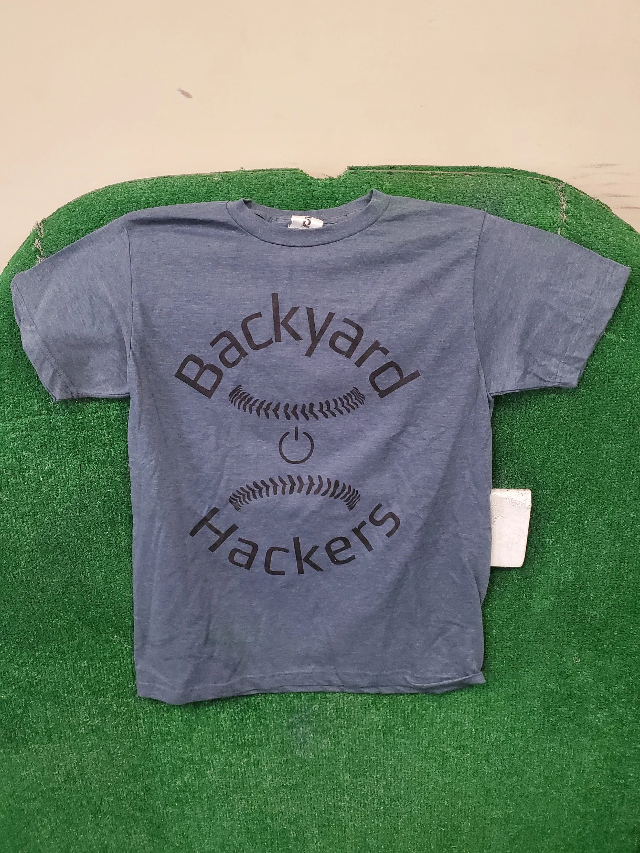 Gray T-shirt with the words 'Backyard Hackers' and a smiley face printed on it, laid on a green textured surface.