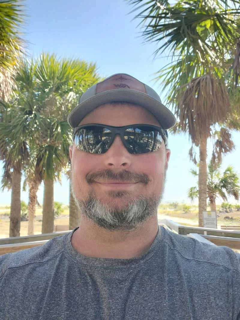Smiling man wearing sunglasses and a cap outdoors with palm trees and a clear blue sky in the background.