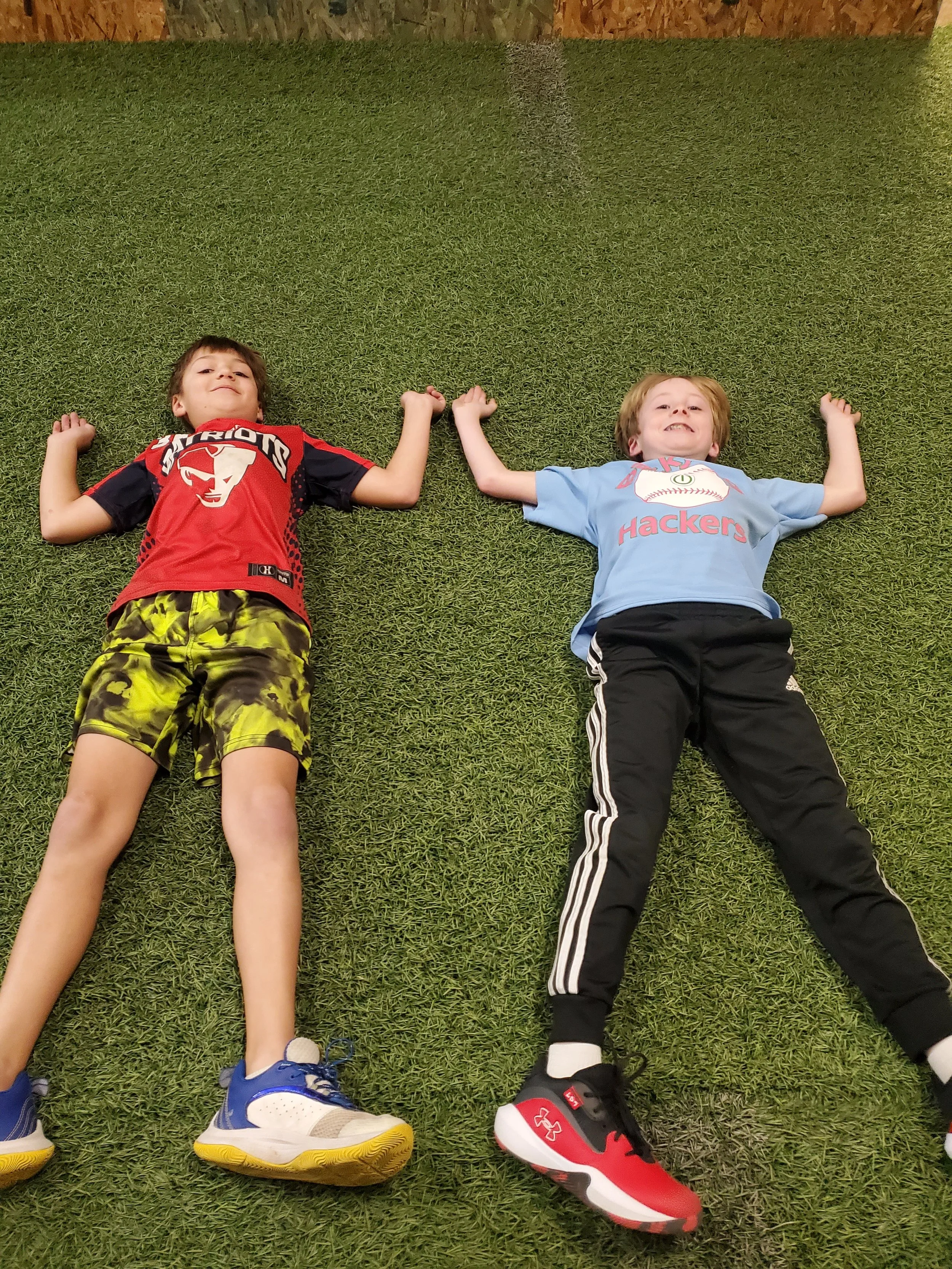Two boys lying on artificial green grass, holding hands above their heads. One boy wears a red sports jersey with a logo and camouflage shorts, blue and white sneakers. The other boy wears a light blue t-shirt with a graphic and black Adidas pants with white stripes, red and black sneakers.