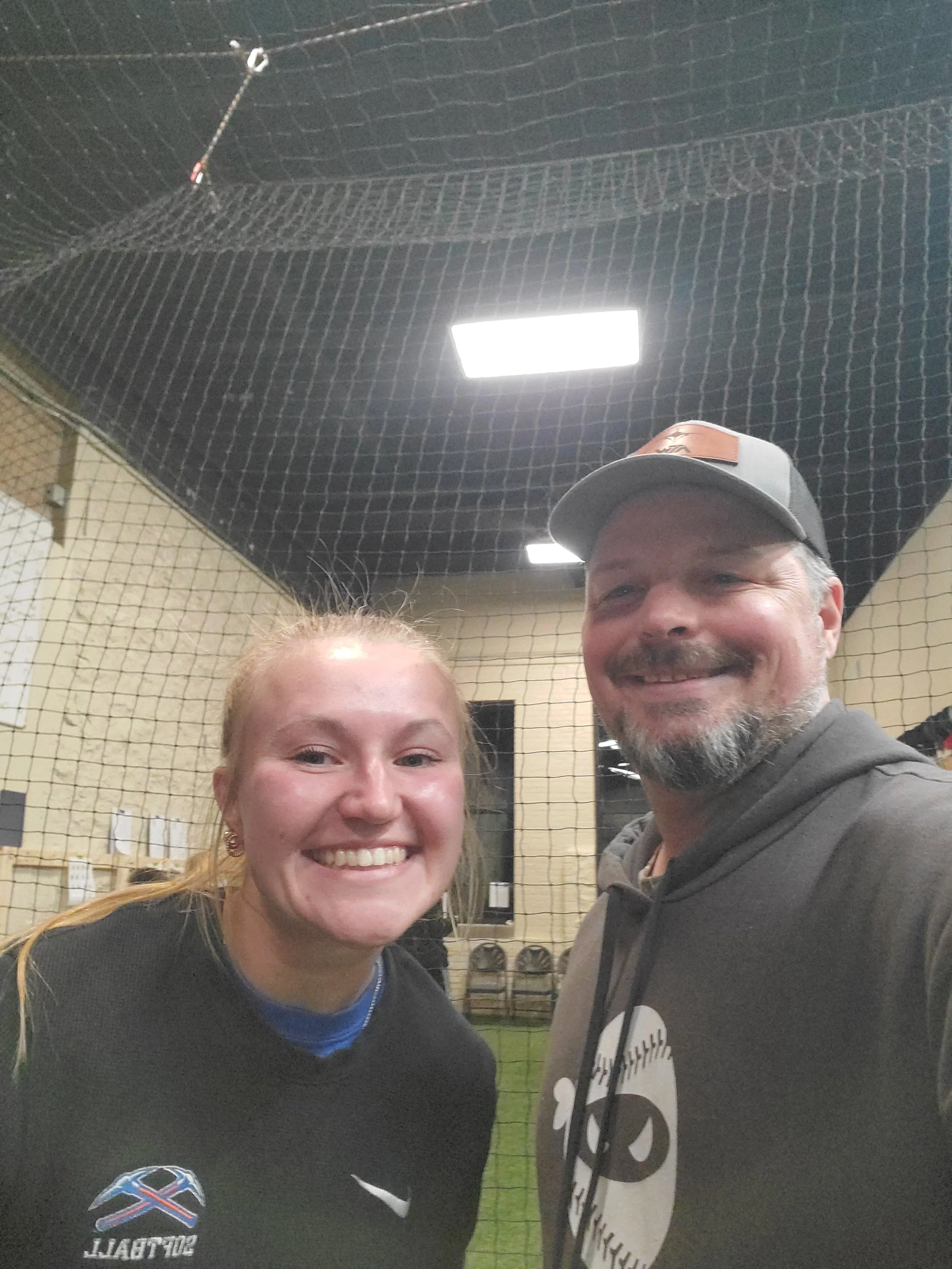 A smiling young  softball player and a man are taking a selfie in front of a sports net inside a gym. The woman has long blonde hair, and the man has a beard and is wearing a gray baseball cap and gray hoodie.
