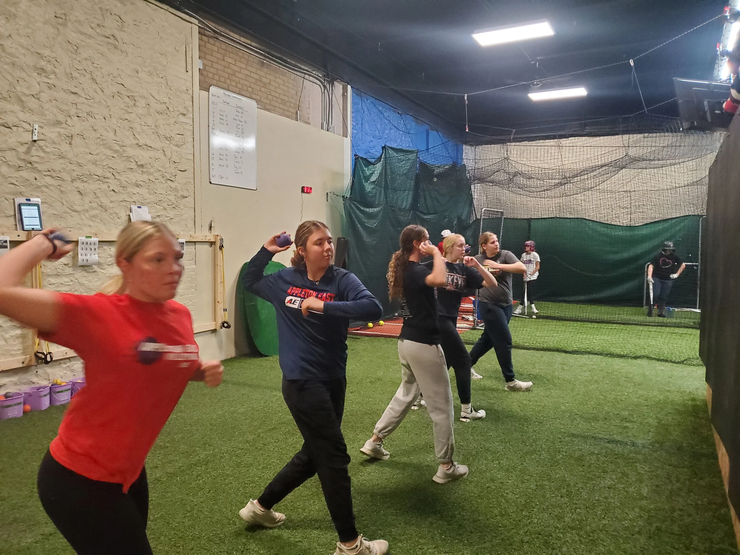 A group of young women practicing softball indoors, working on their throwing velocity with plyoballs, with a netted area and a whiteboard in the background.