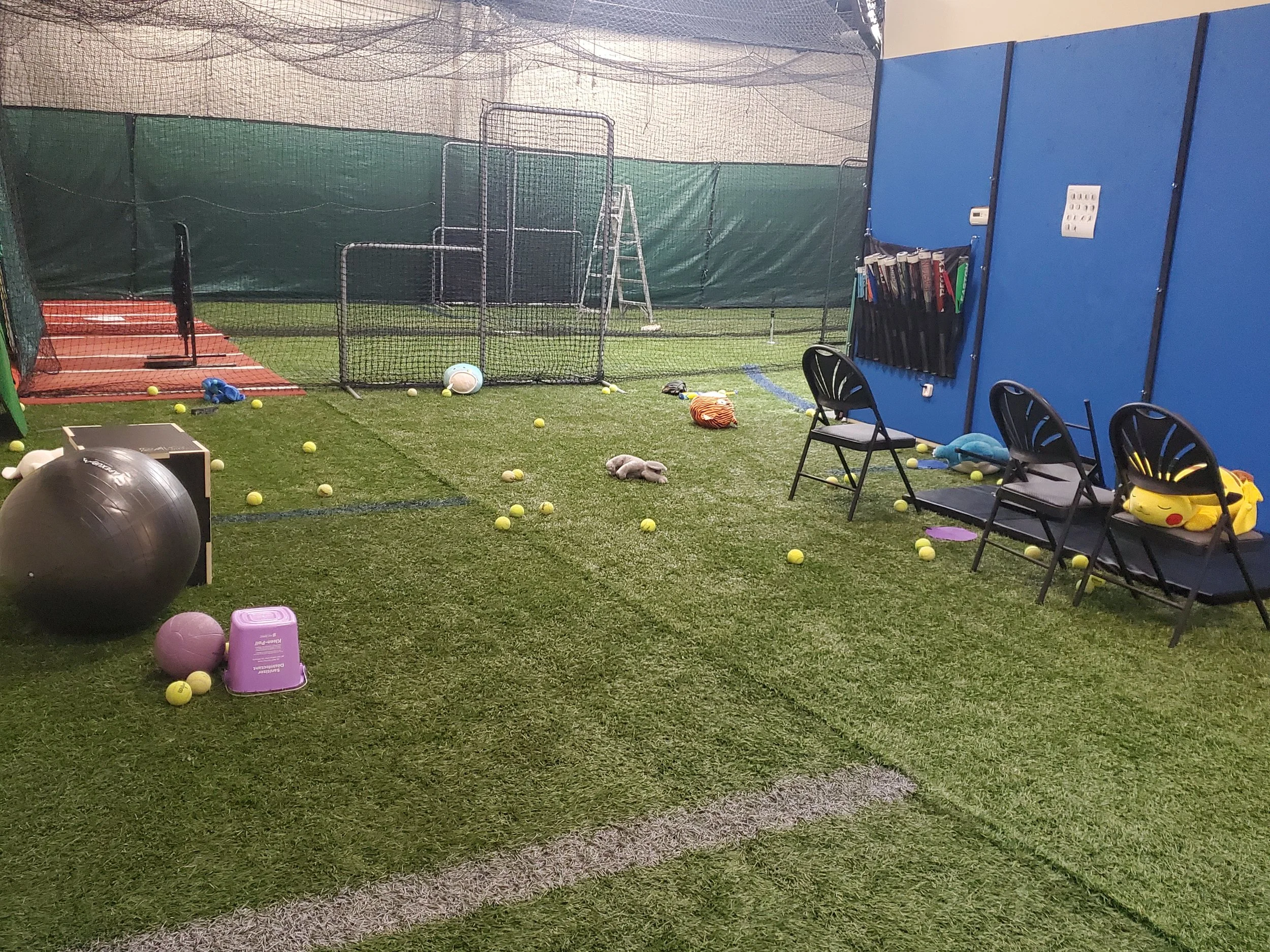 Indoor sports practice area with a batting cage, tennis balls scattered across artificial grass, chairs, and various sports equipment including a golf ball, a large exercise ball, and a basket with tennis rackets, against a blue and white wall.