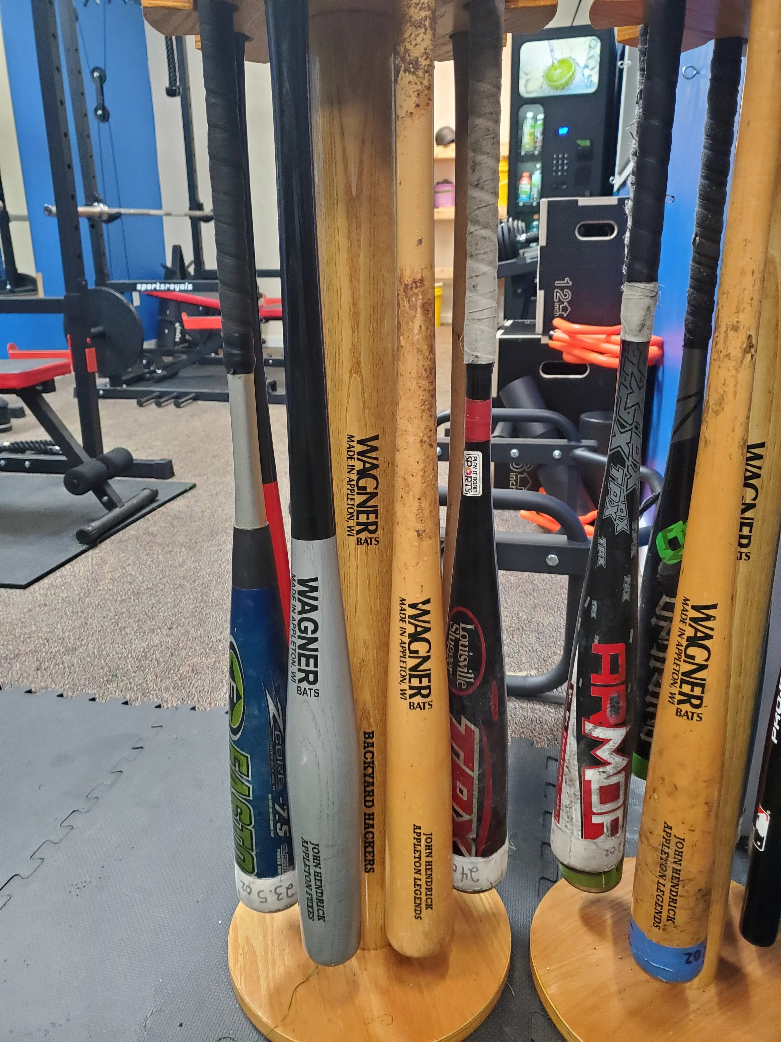A display of various baseball bats with different brands and designs, standing vertically on a wooden stand in a gym or sports equipment store.