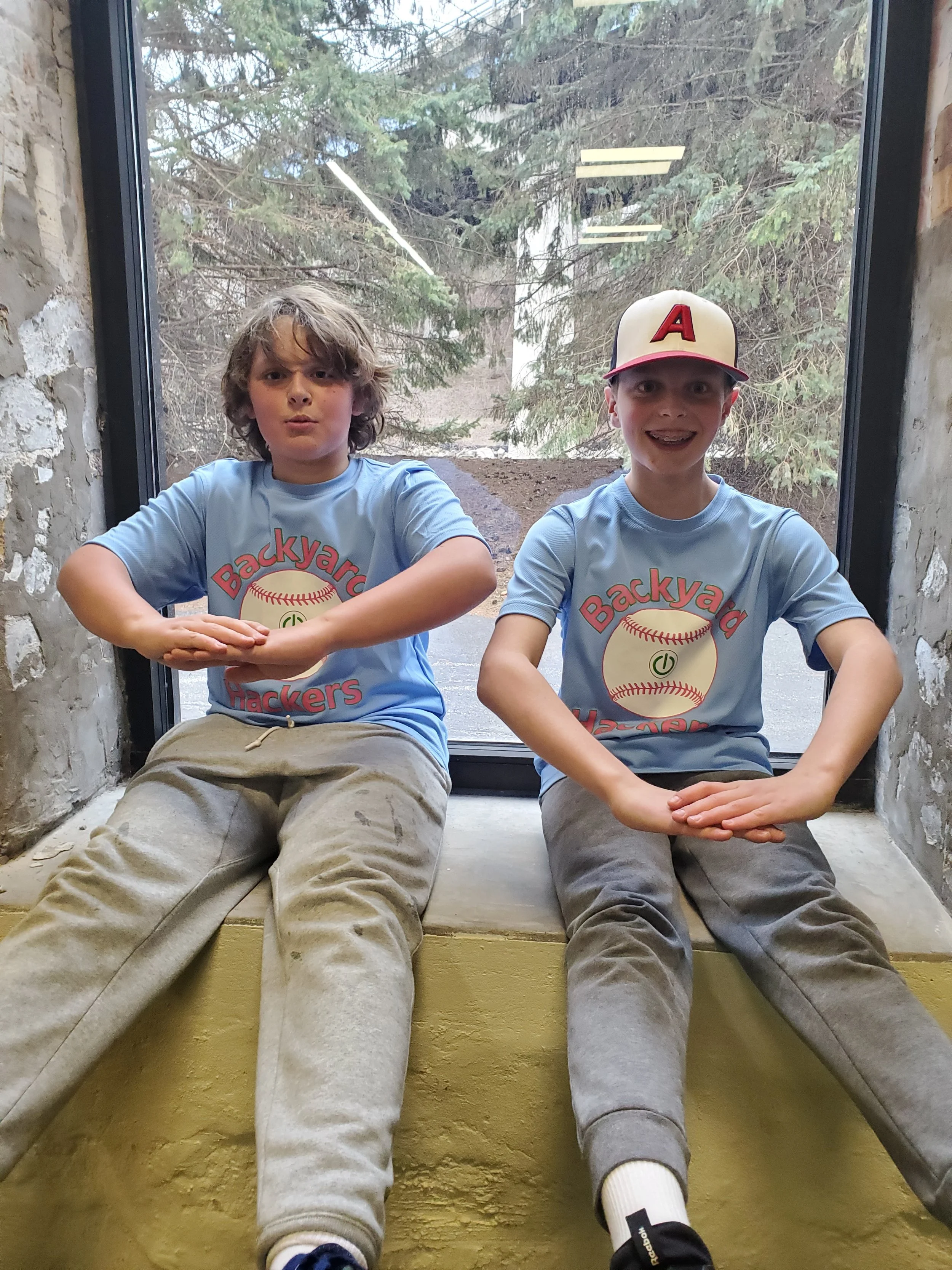 Two boys sitting on a ledge in front of a large window with a forested background. They are wearing matching light blue t-shirts with a baseball graphic and the words 'Backyard Hackers'. One boy has curly brown hair and the other is wearing a baseball cap with an 'A' on it. Both have their arms crossed or resting on their legs, looking at the camera.