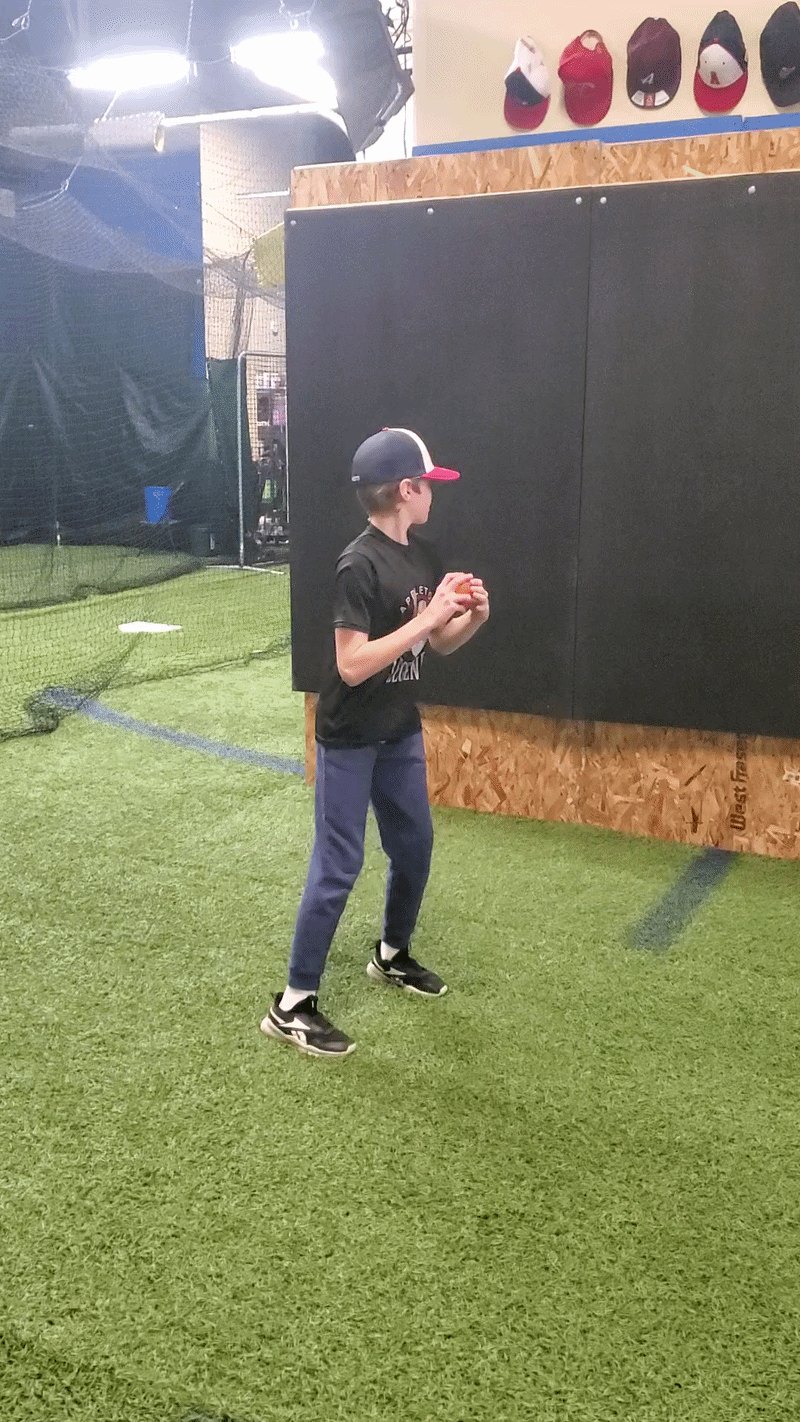 A young boy in a baseball cap holding a baseball at an indoor sports facility with artificial turf, black padded walls, and a basketball and hats displayed on the wall.