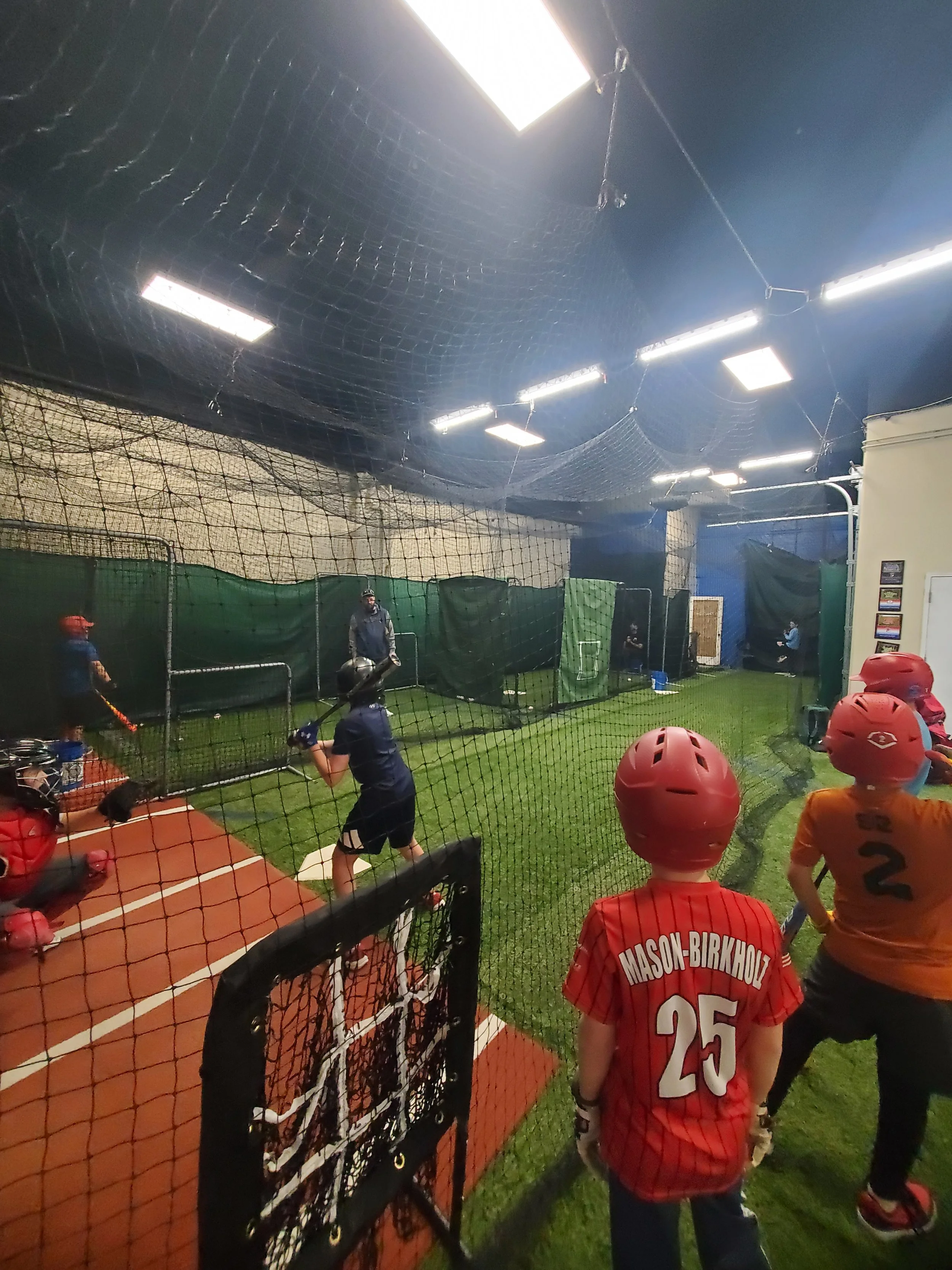 Indoor baseball training facility with players practicing batting, wearing helmets and jerseys, some with names and numbers, behind a protective net.