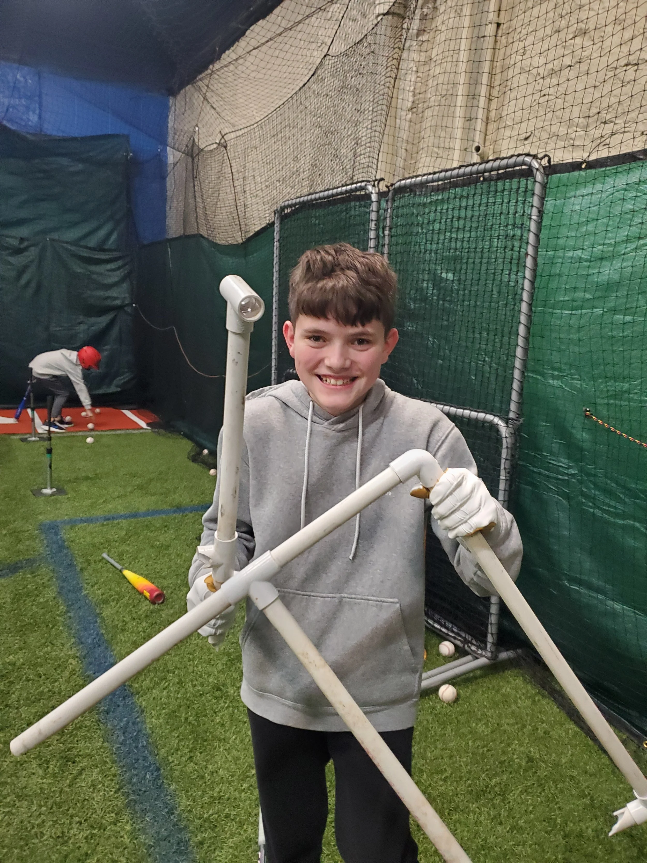 A young boy smiling and holding a structure made of white PVC pipes indoors. In the background, a person in a red helmet is practicing golf, and there are various sports nets and equipment around.