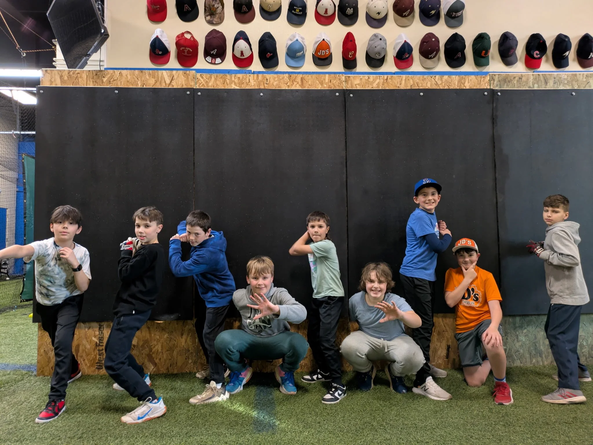 A group of children posing inside a sports facility in front of a black padded wall and a wooden panel, with numerous baseball caps displayed on the wall above them.