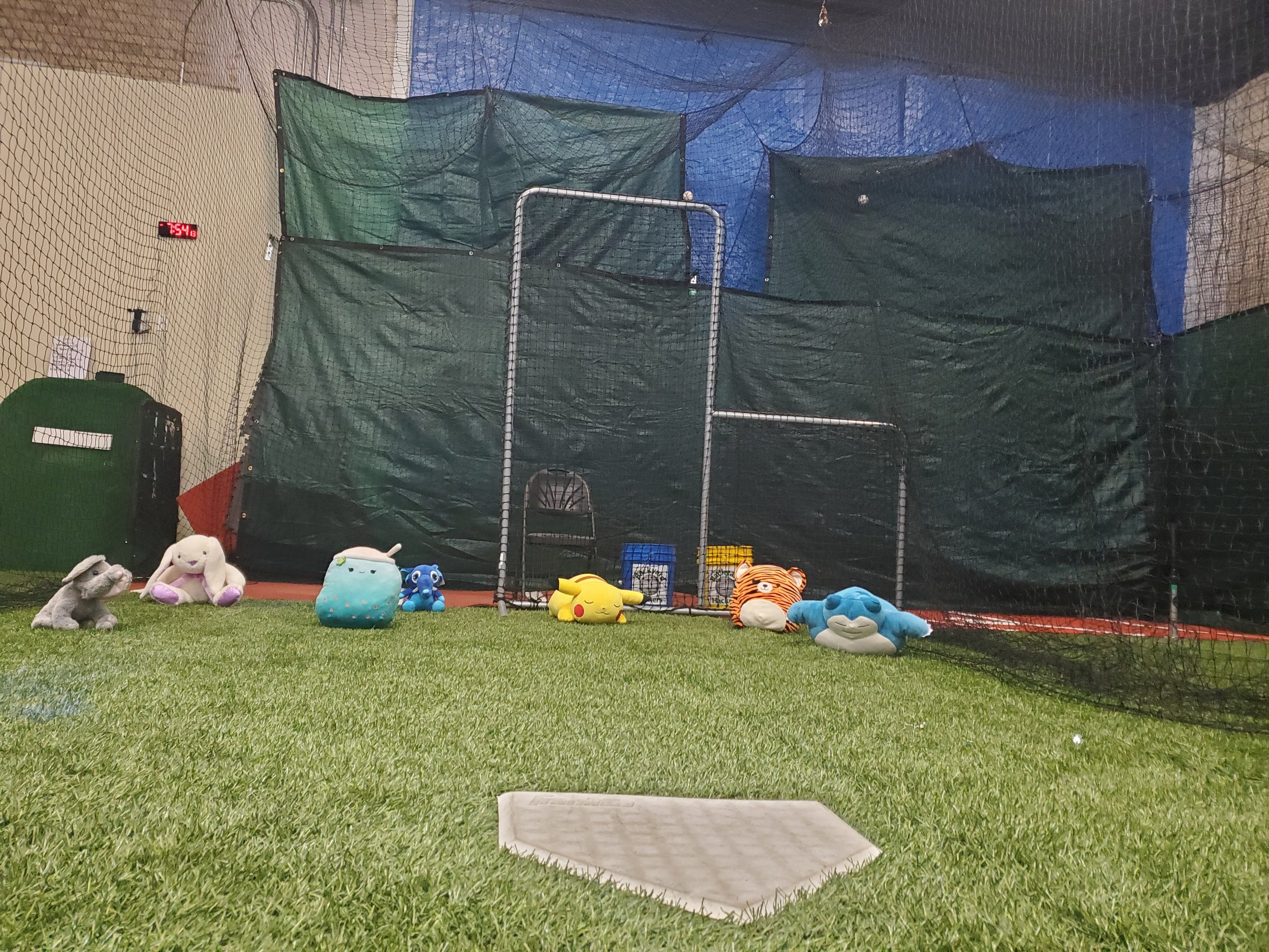 A batting cage at an indoor baseball or softball facility, with plush toys arranged on the green artificial turf in front. The cage is enclosed with netting, and there is a scoreboard showing a score of -754. The background features blue walls and additional netting.
