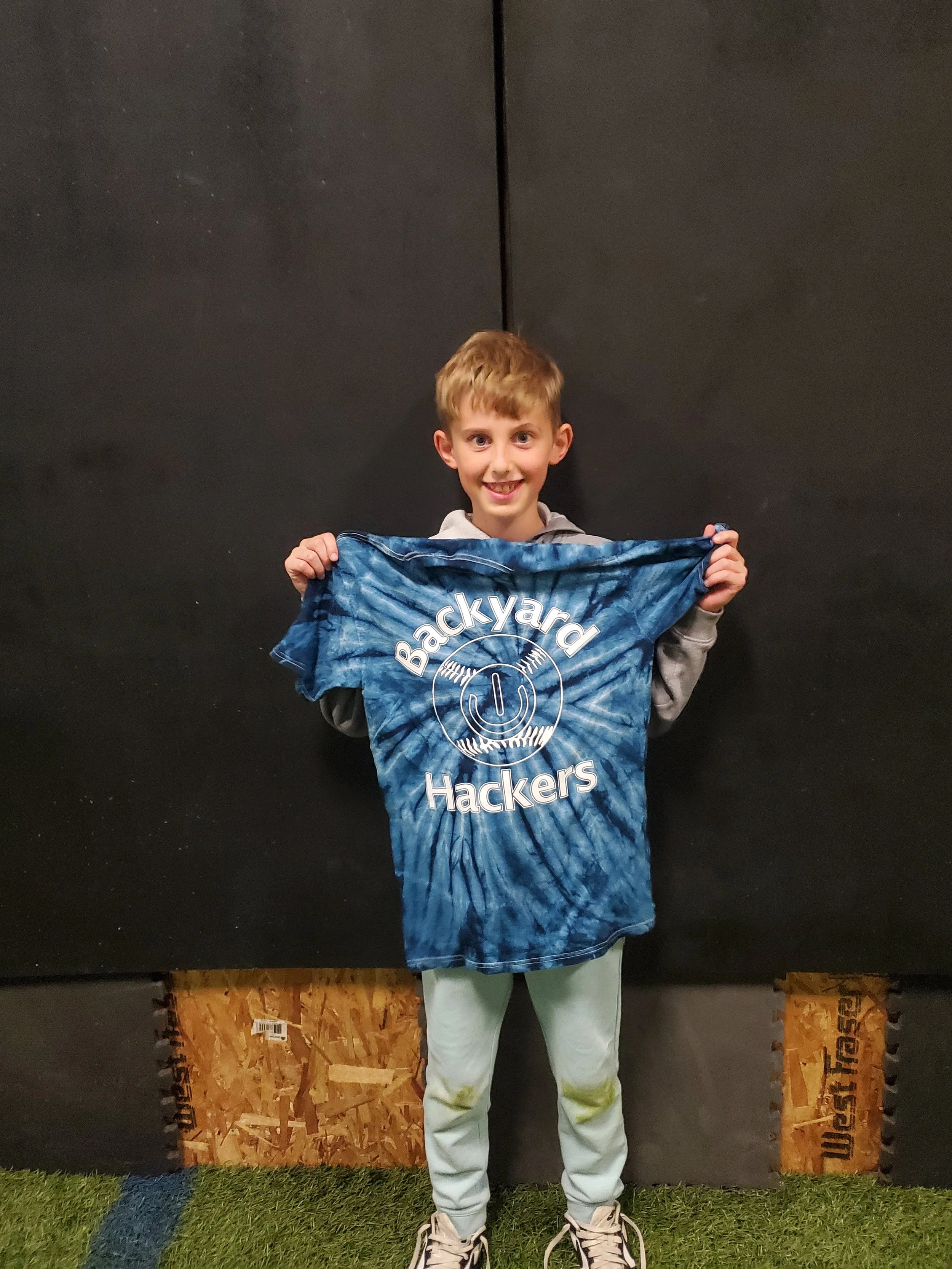 A young boy smiling and holding a blue t-shirt with the words 'Backyard Hackers' and a headset graphic, standing in front of a black wall and green turf ground.