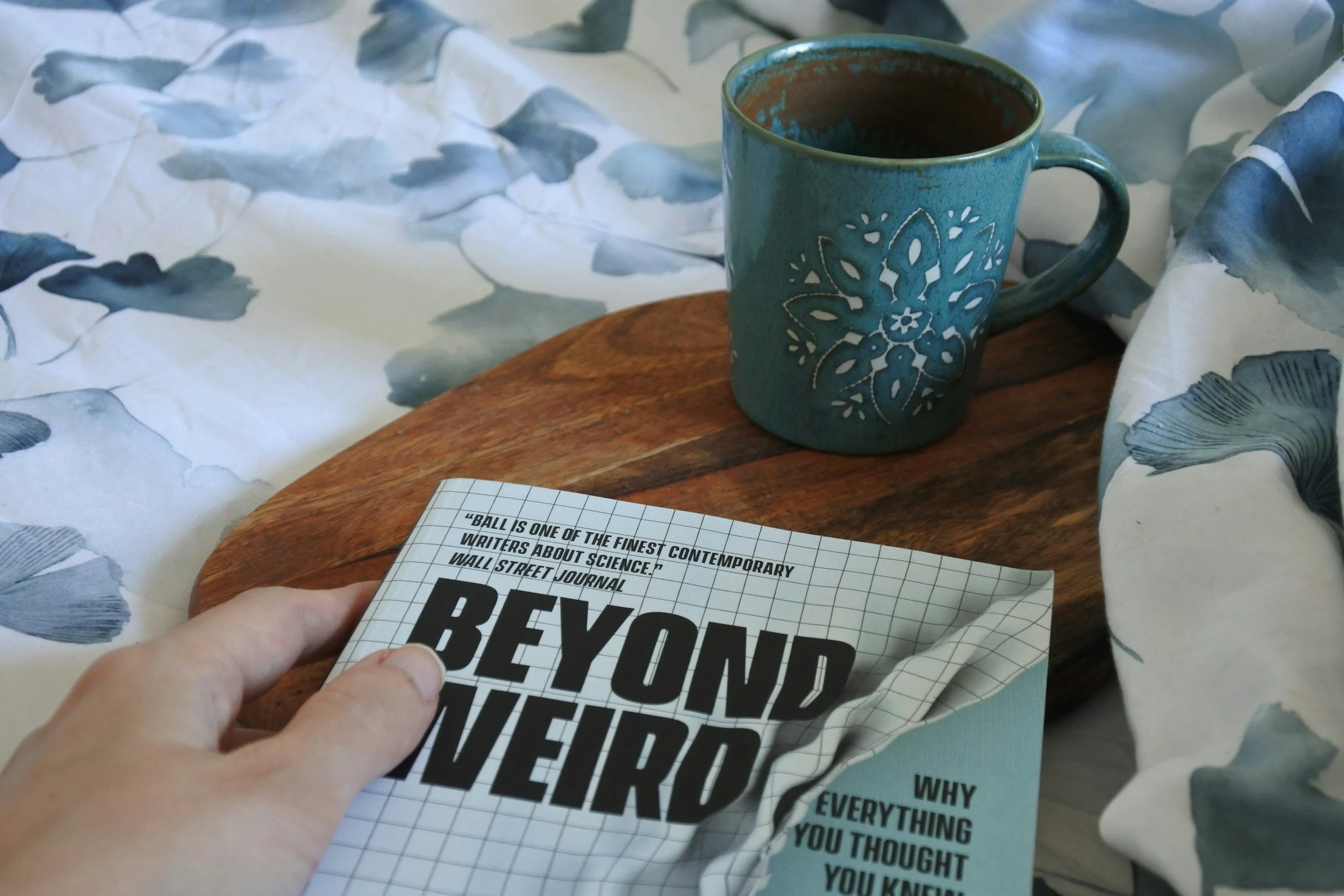 A blue ceramic mug with a decorative hole pattern, filled with a dark beverage, on a wooden surface surrounded by crumpled paper with a partial view of a person's hand holding a motivational printed paper.