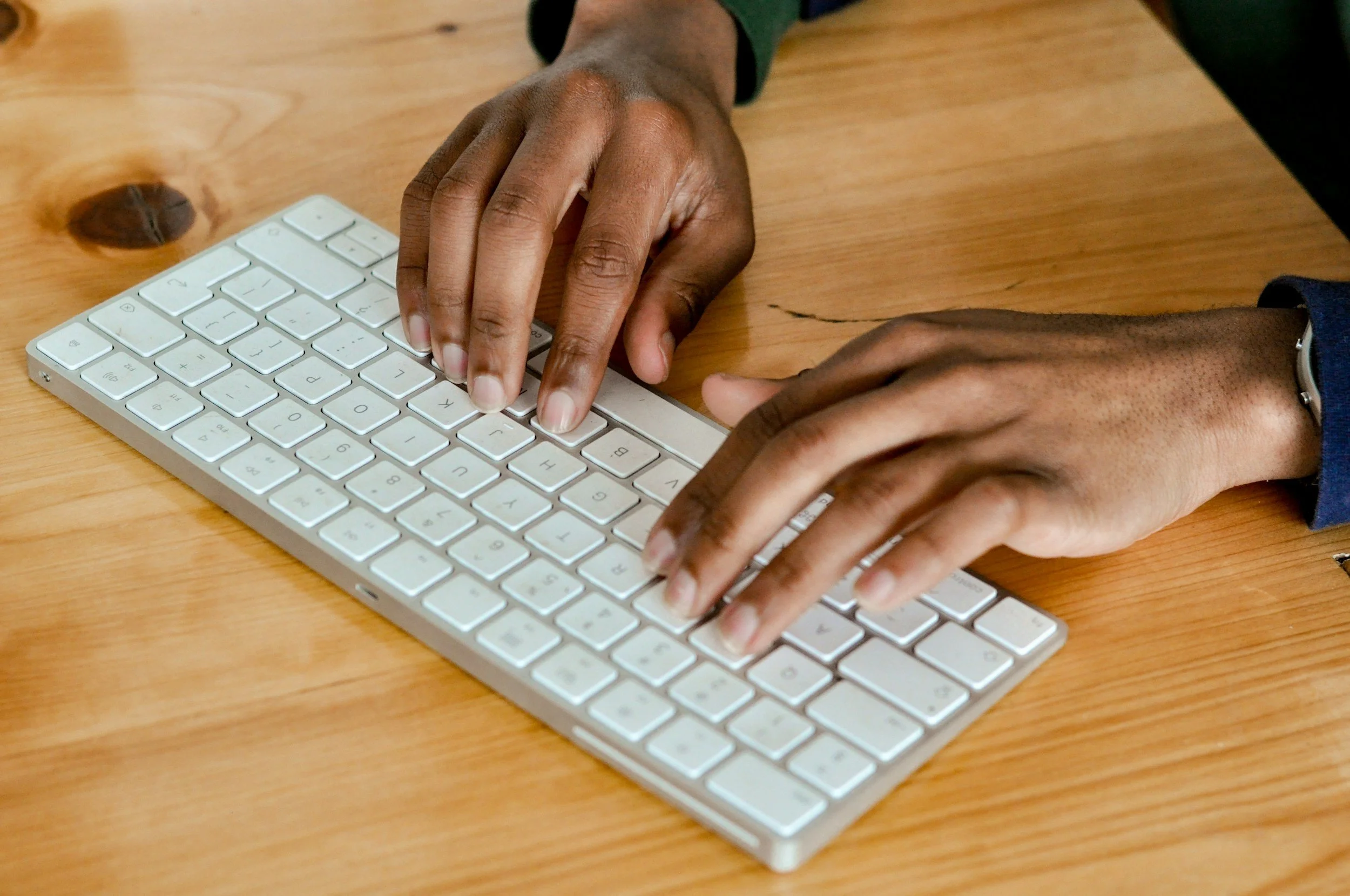 Two hands typing on a white computer keyboard placed on a wooden table.