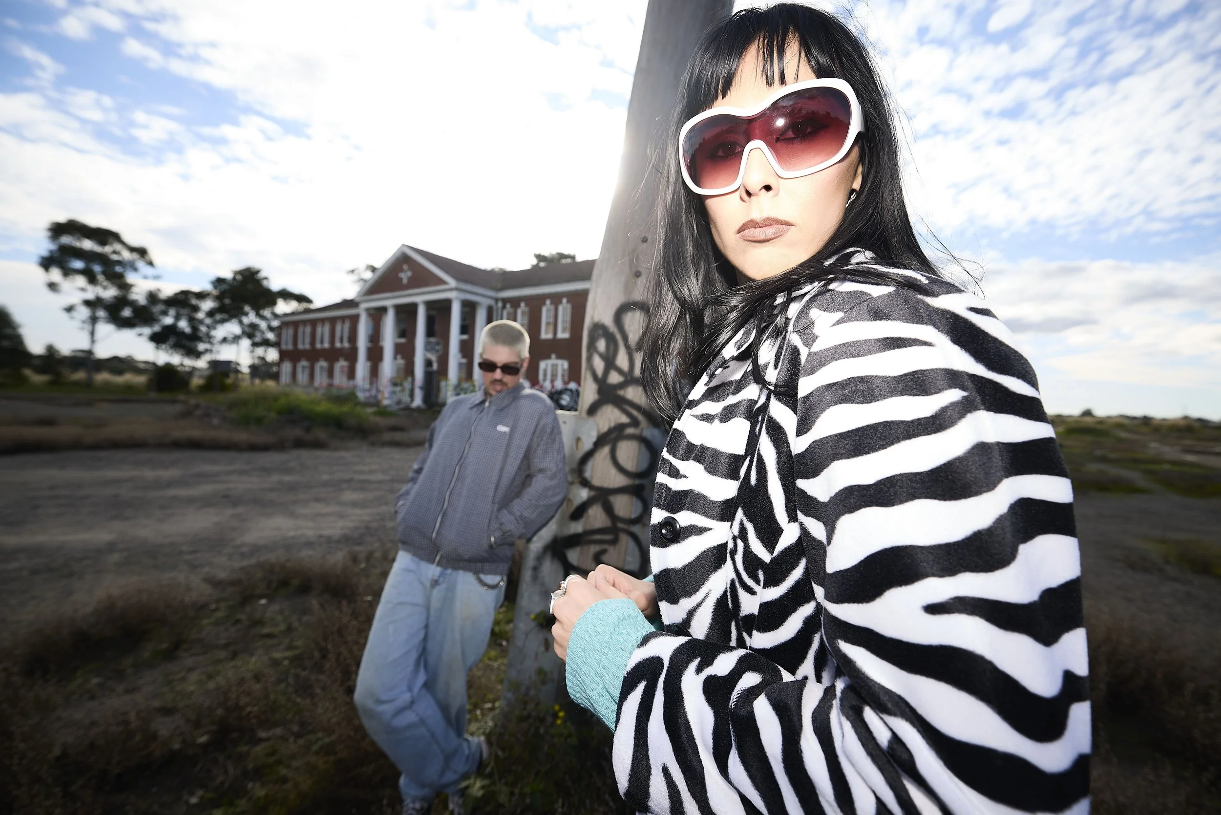 Lahgo. A woman with black hair wearing large sunglasses and a black-and-white striped blazer stands outdoors next to a tree. A man wearing sunglasses and a gray hoodie stands further back near a historic brick building with white columns.